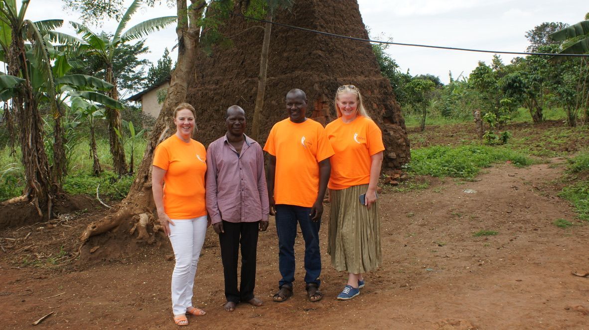 Four people stand together in front of a tall, brown structure outdoors; two wear orange shirts, one wears a patterned shirt.
