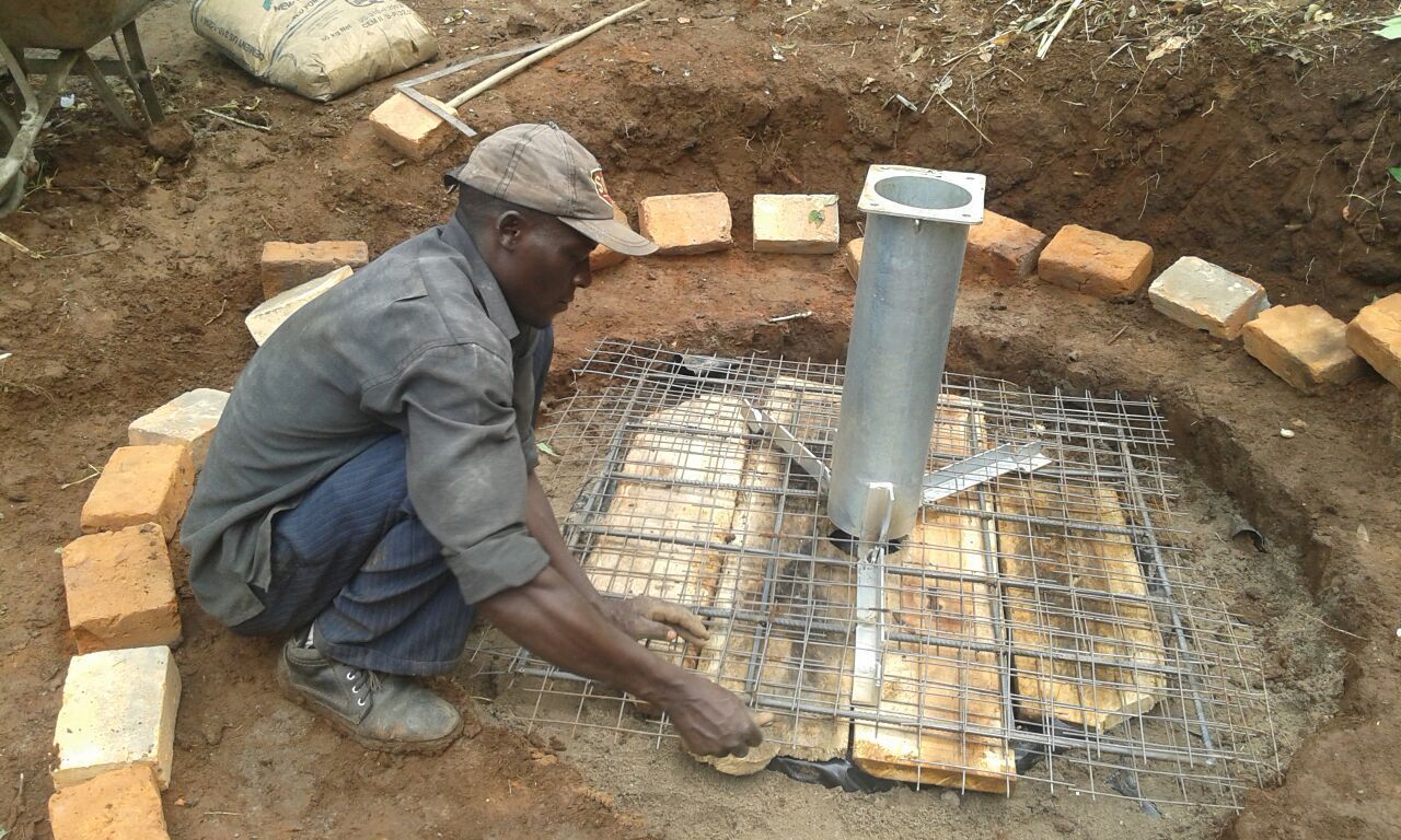 Man installing metal grate over wooden pieces inside a circular brick structure; metal tube in the center.