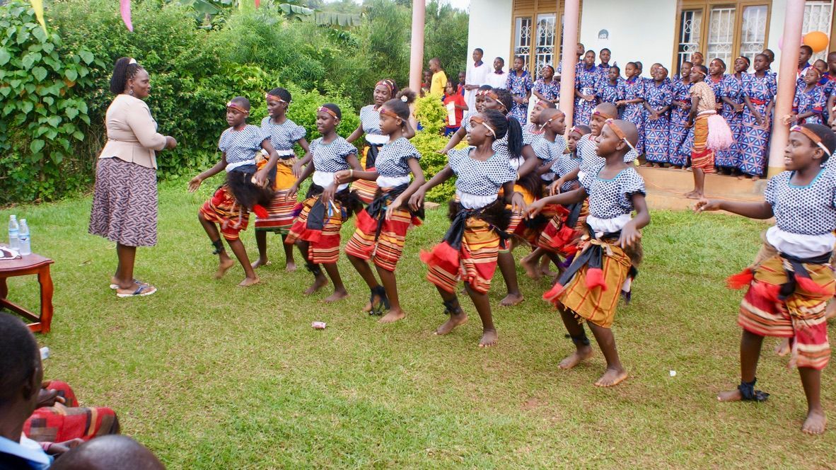 Children in colorful skirts dance on grass, watched by an adult. A building and other people are in the background.
