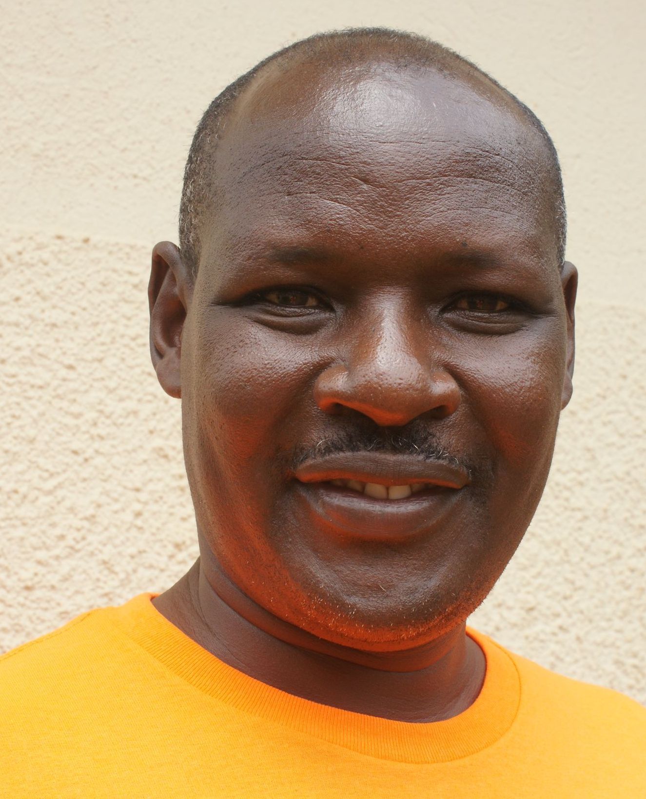 A man with dark skin wearing an orange shirt is smiling, looking directly at the camera.