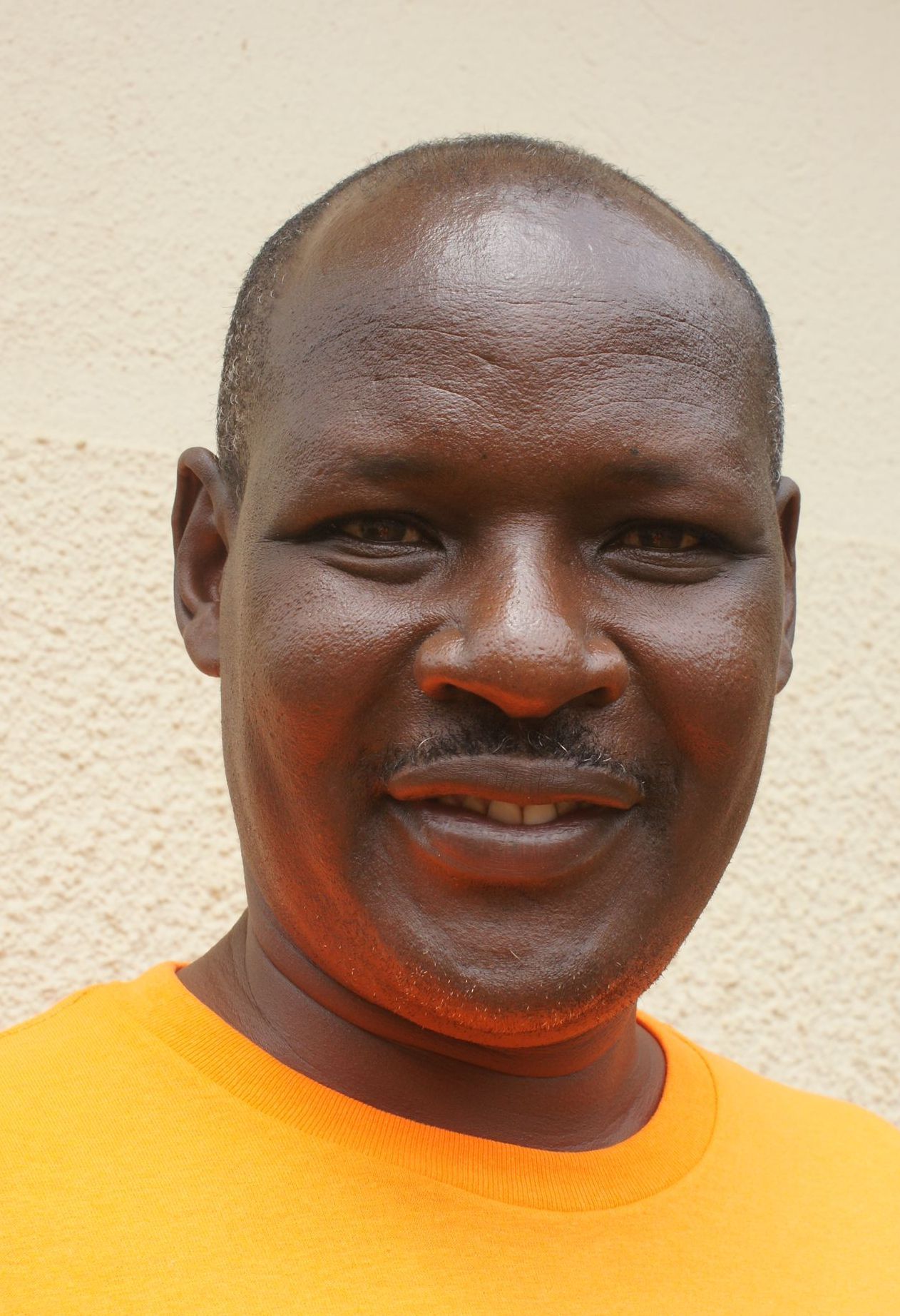 Man with dark skin wearing an orange shirt, smiling in front of a light-colored wall.
