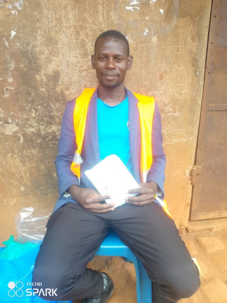Man seated on blue stool, wearing yellow vest, blue shirt, and blazer, holding papers. Brown wall background.