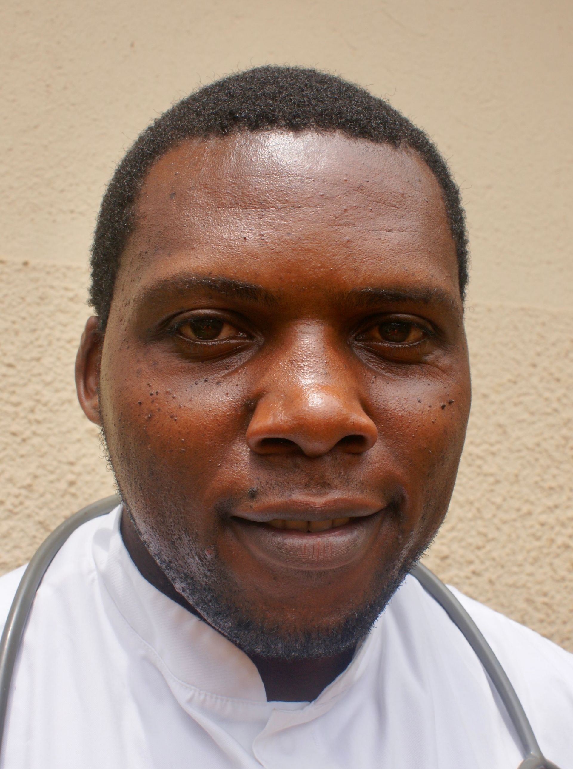 Man wearing a white coat and stethoscope, looking at the camera. Neutral expression. Beige background.