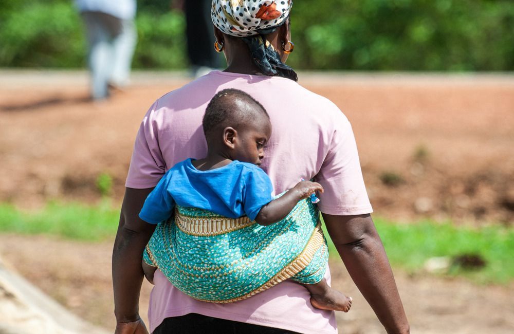 Woman carrying a child on her back; child in a blue shirt, teal carrier. Outdoors, walking.