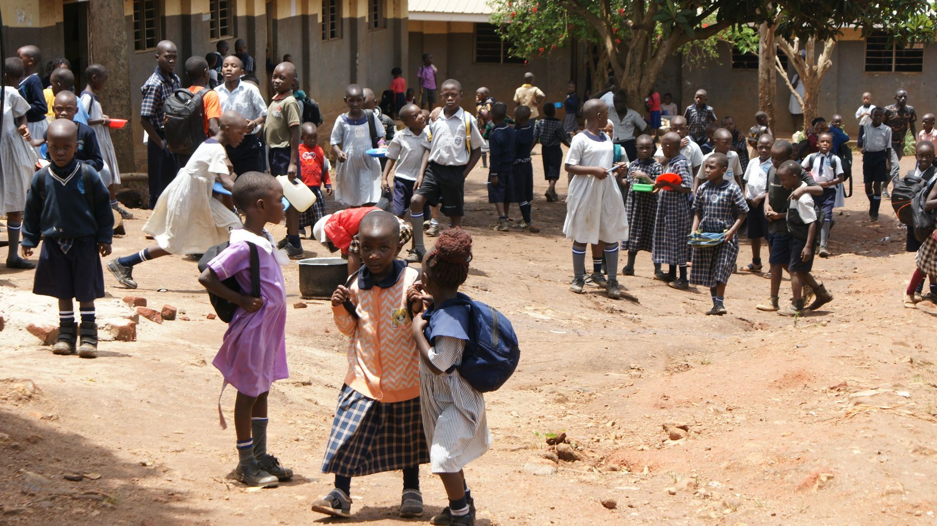 School children in uniform milling about a dusty outdoor area, some with backpacks.