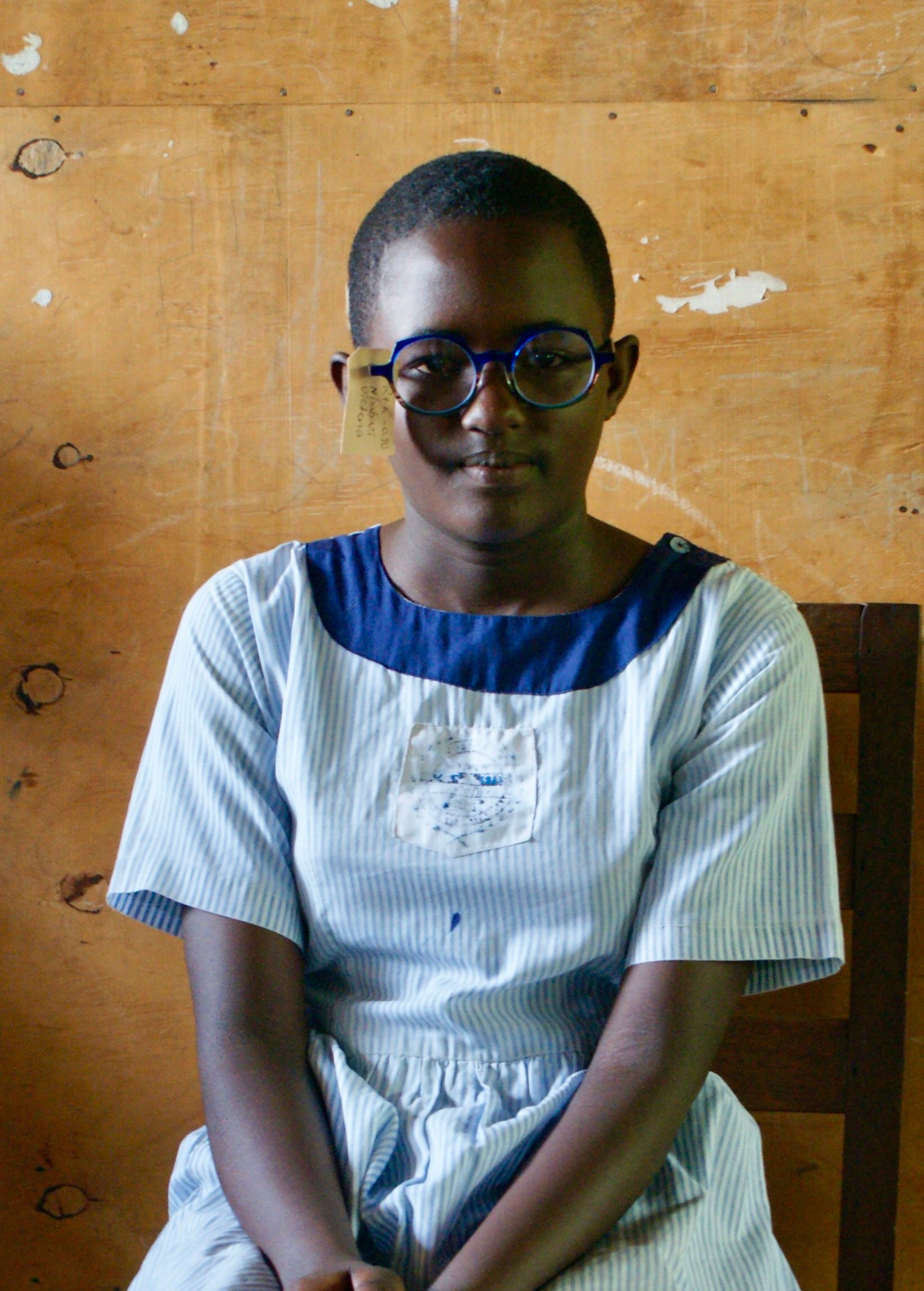 Young person wearing glasses and a blue-collared dress, sitting in front of a wooden wall.