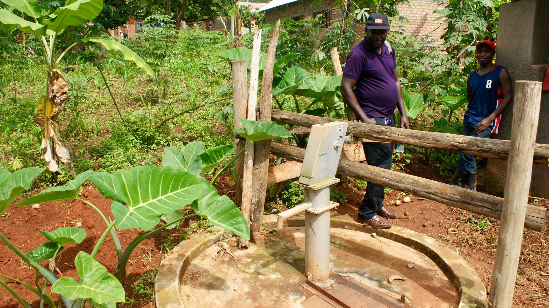 Two men by a well with a wooden fence in a lush, green environment. One man wears a purple shirt and the other a tank top.