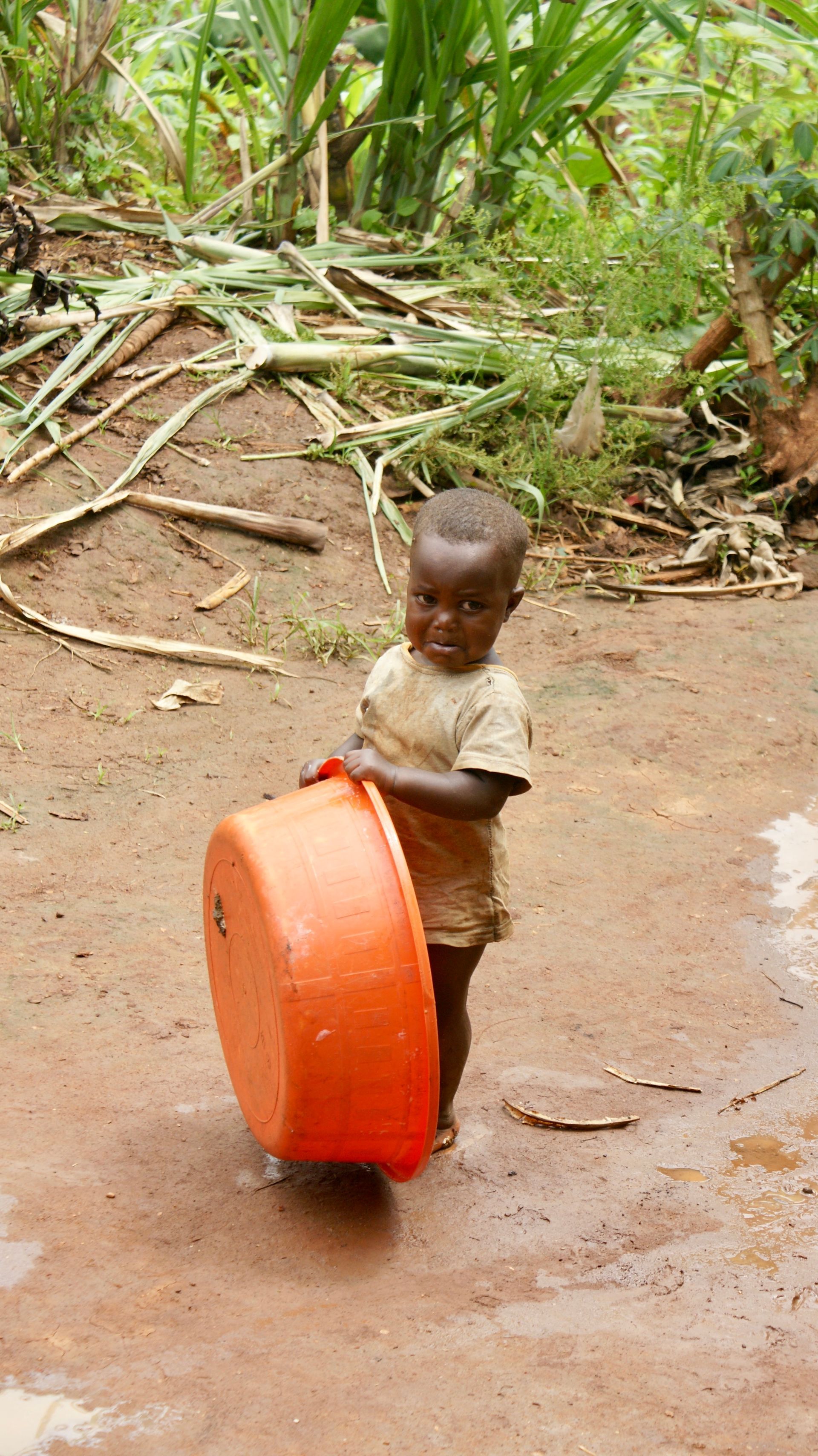 Child holding an orange basin, standing outdoors on muddy ground. Green foliage in the background.