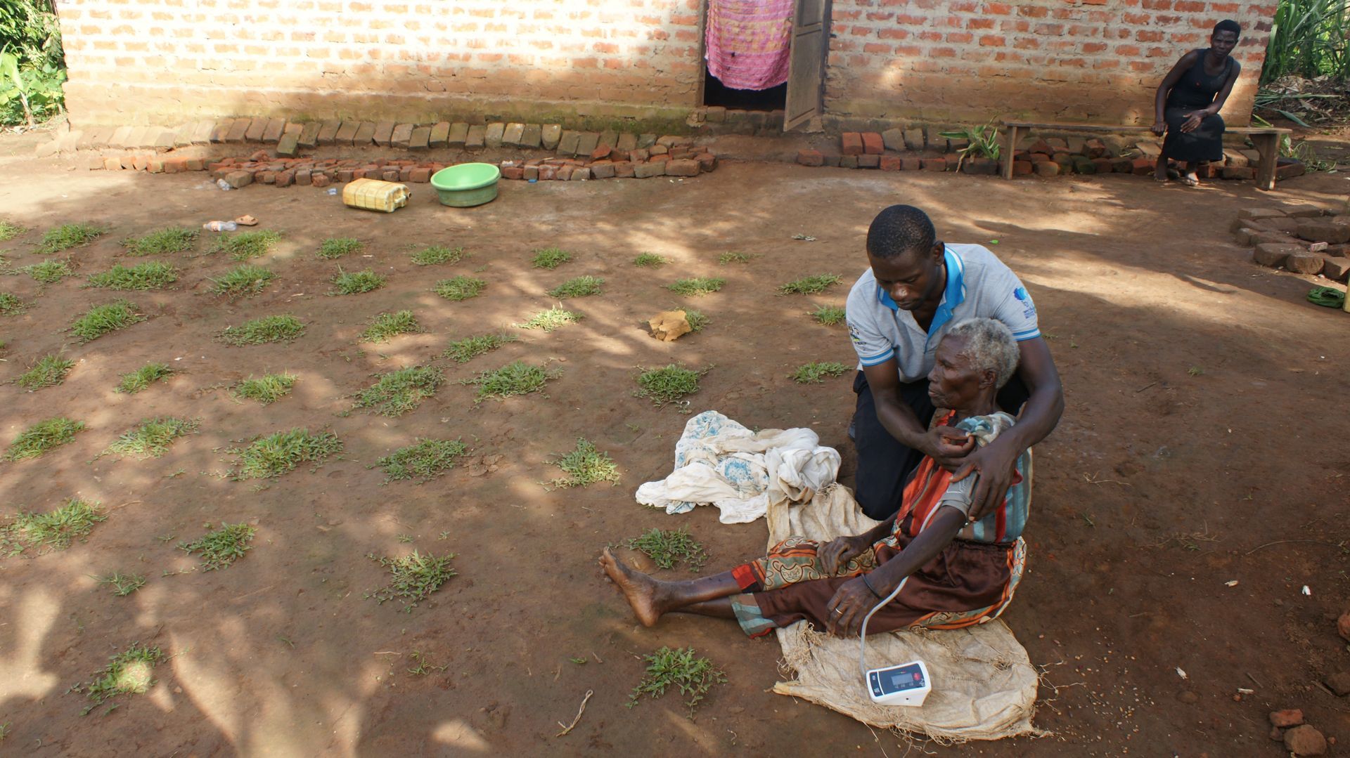 Person helping elderly person outside a brick building. The elderly person sits on the ground.