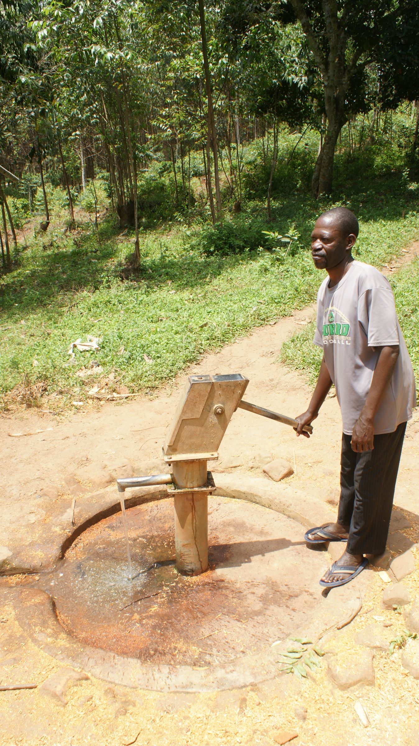 Man pumping water from a wooden well in a grassy outdoor area.