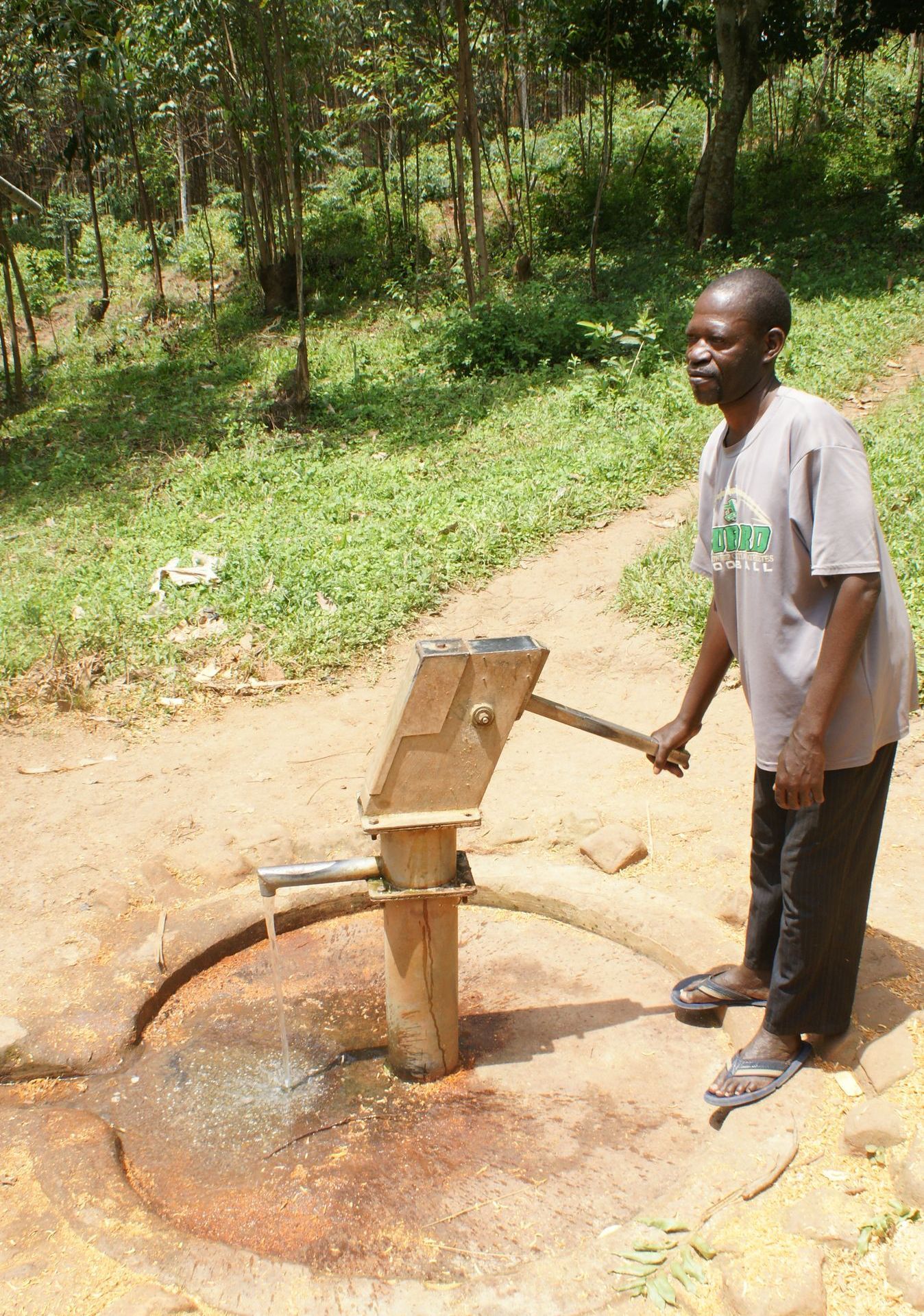 Man pumping water from a well outdoors, with a wooded background.