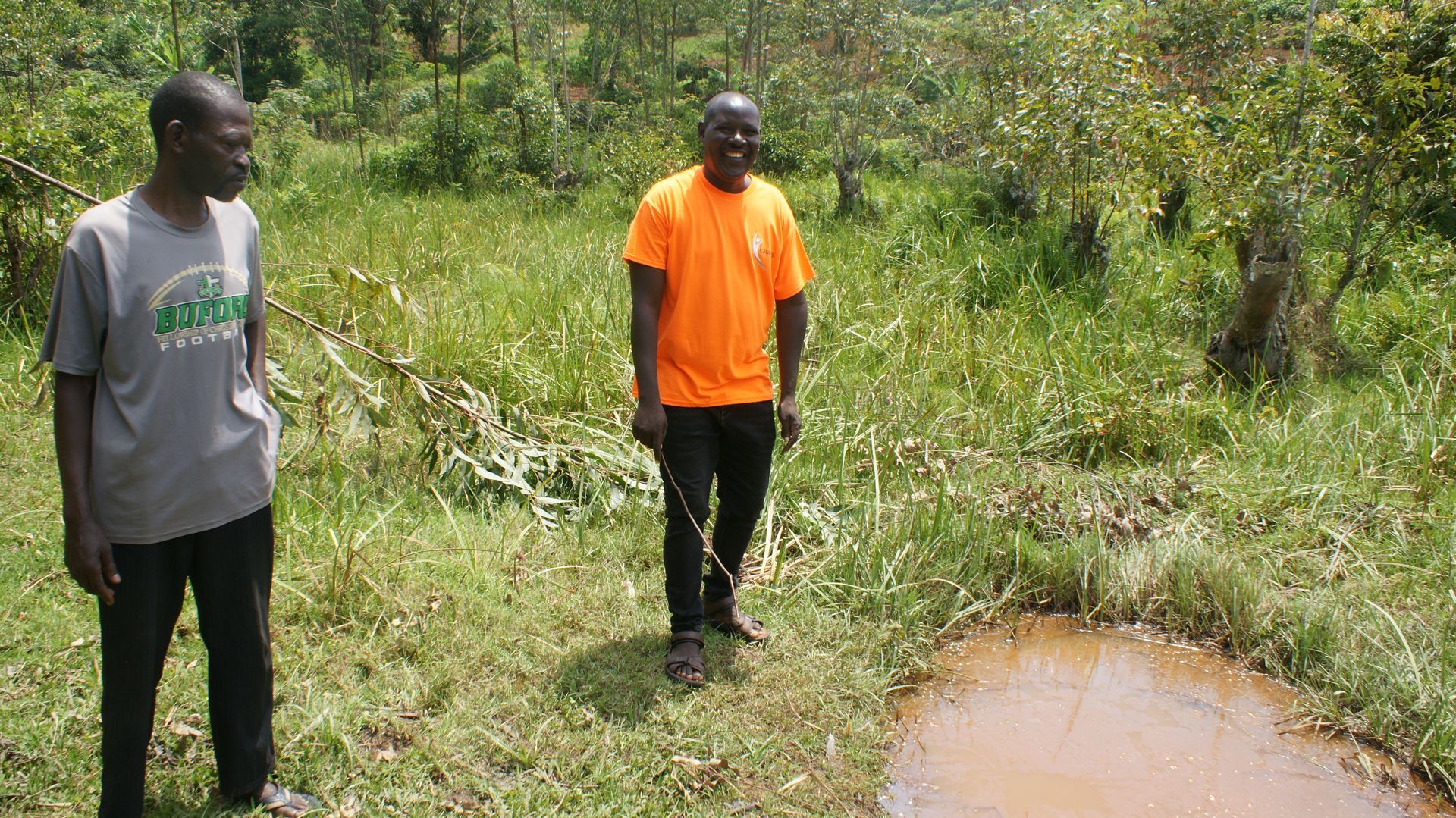 Two men standing in grassy area near a small puddle. Trees in the background.