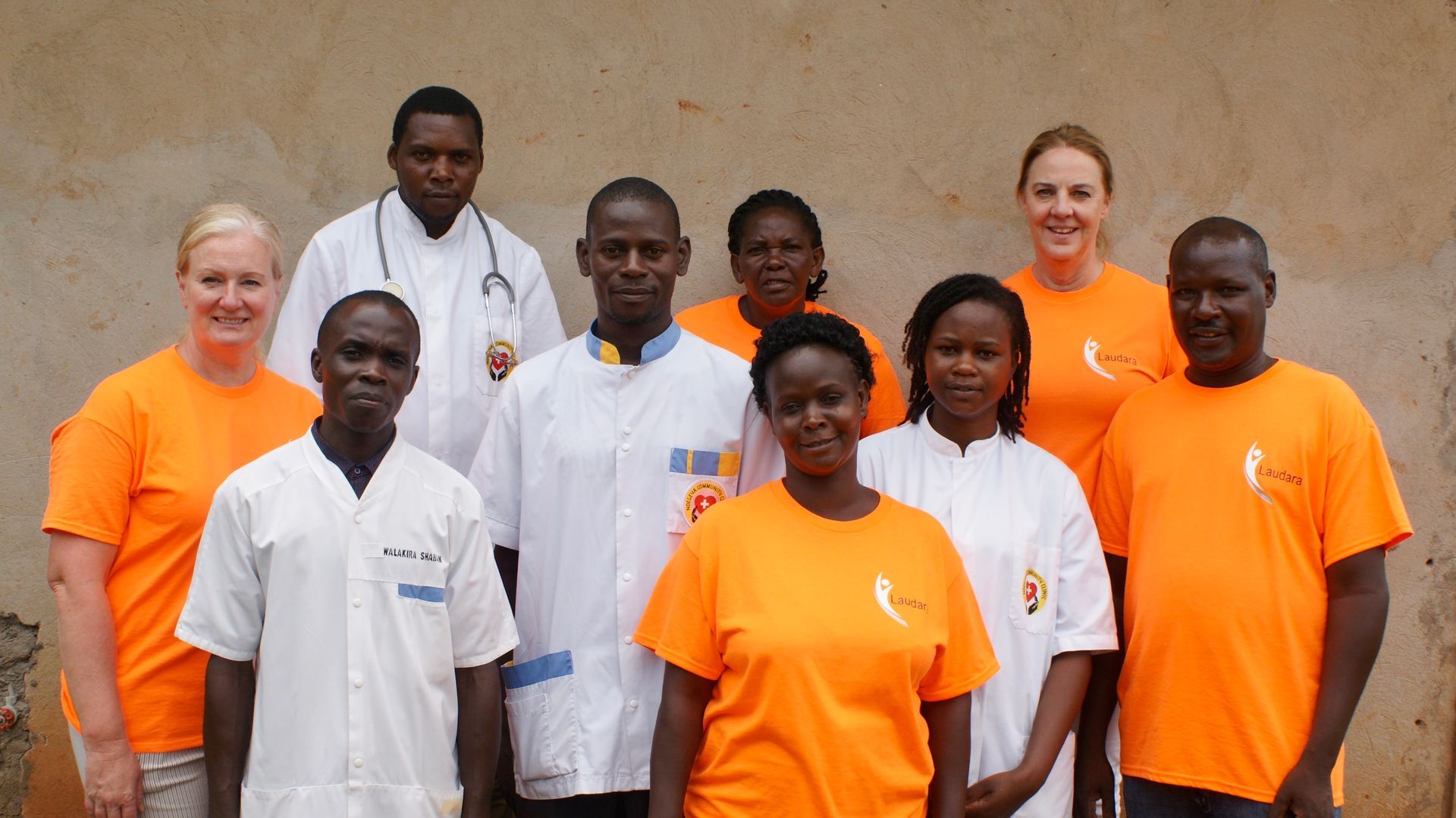Group of health workers in orange shirts and white coats posing together outdoors.