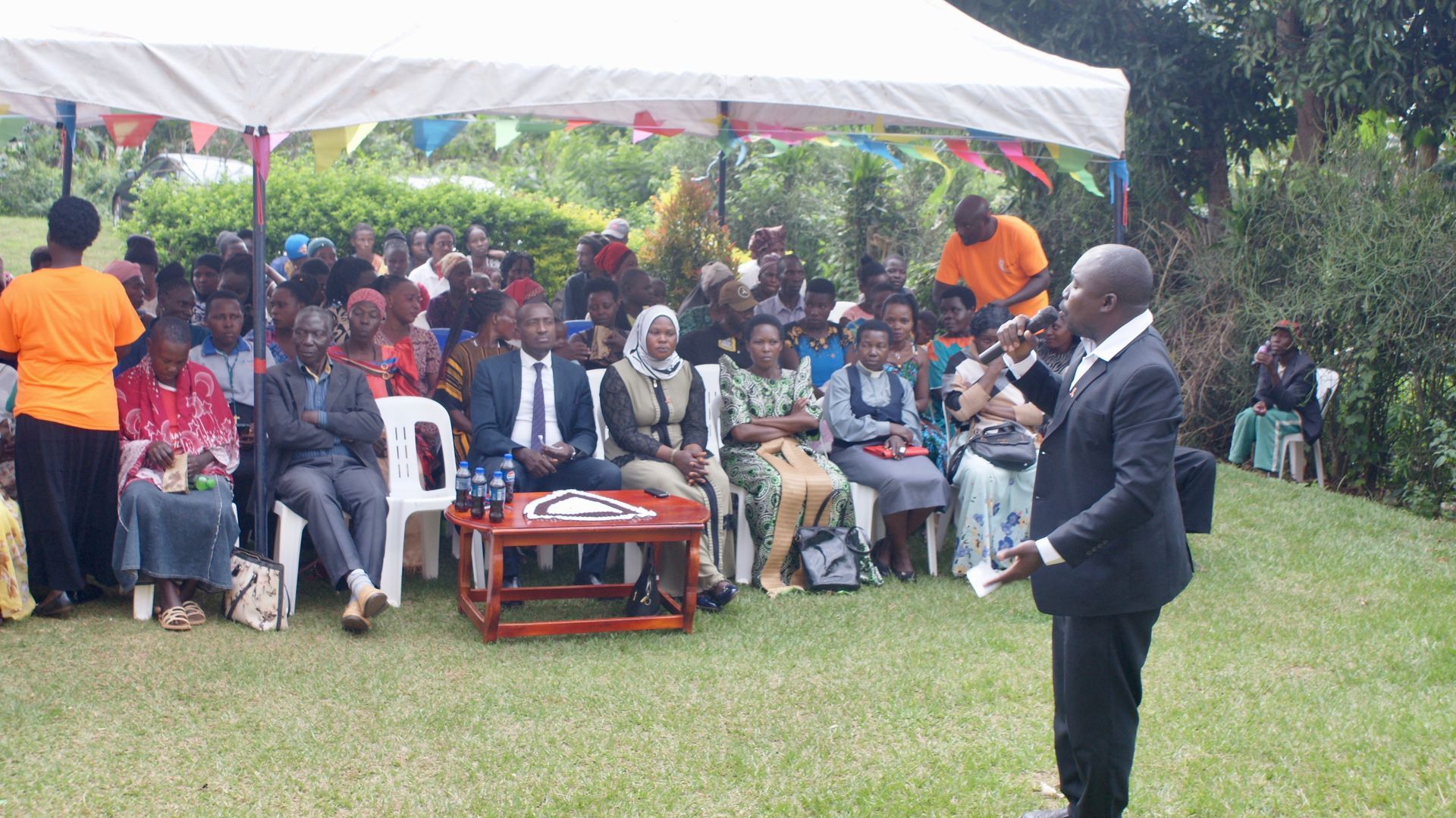Man speaking to a seated crowd under a tent; green grass, colorful decorations.