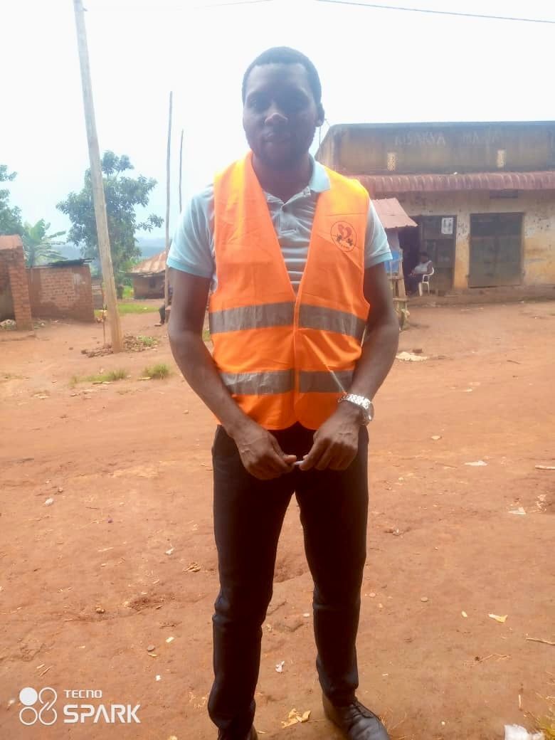 Man in orange vest and blue shirt standing outside a building.