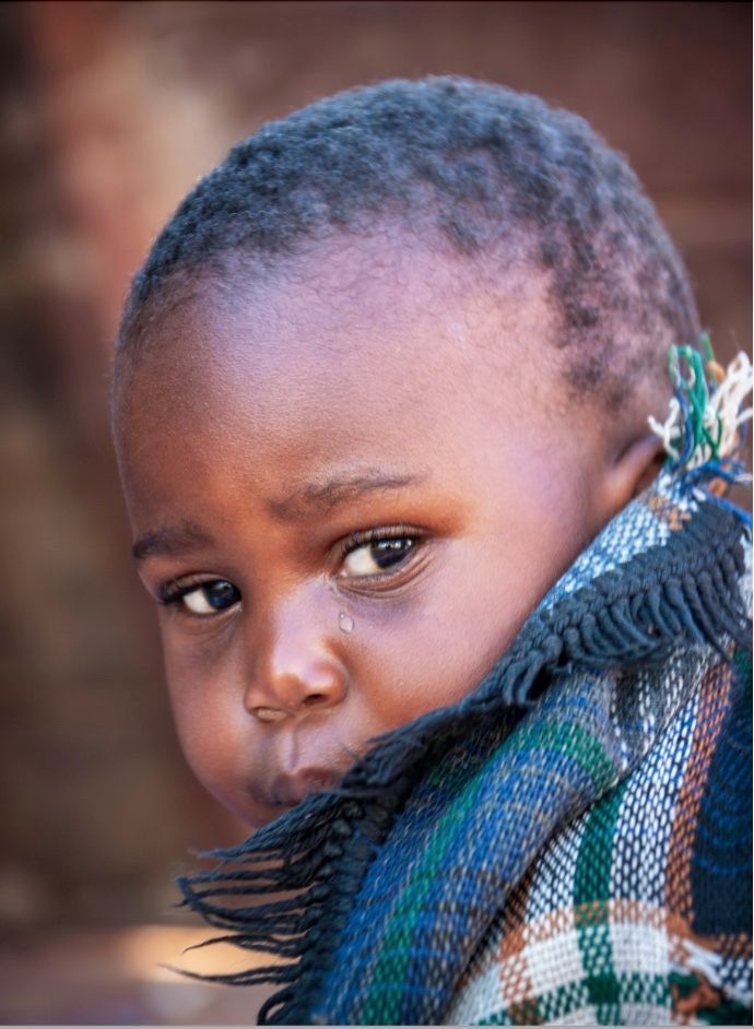 Child wrapped in a patterned blanket, looking somber with a single tear, against a blurred background.