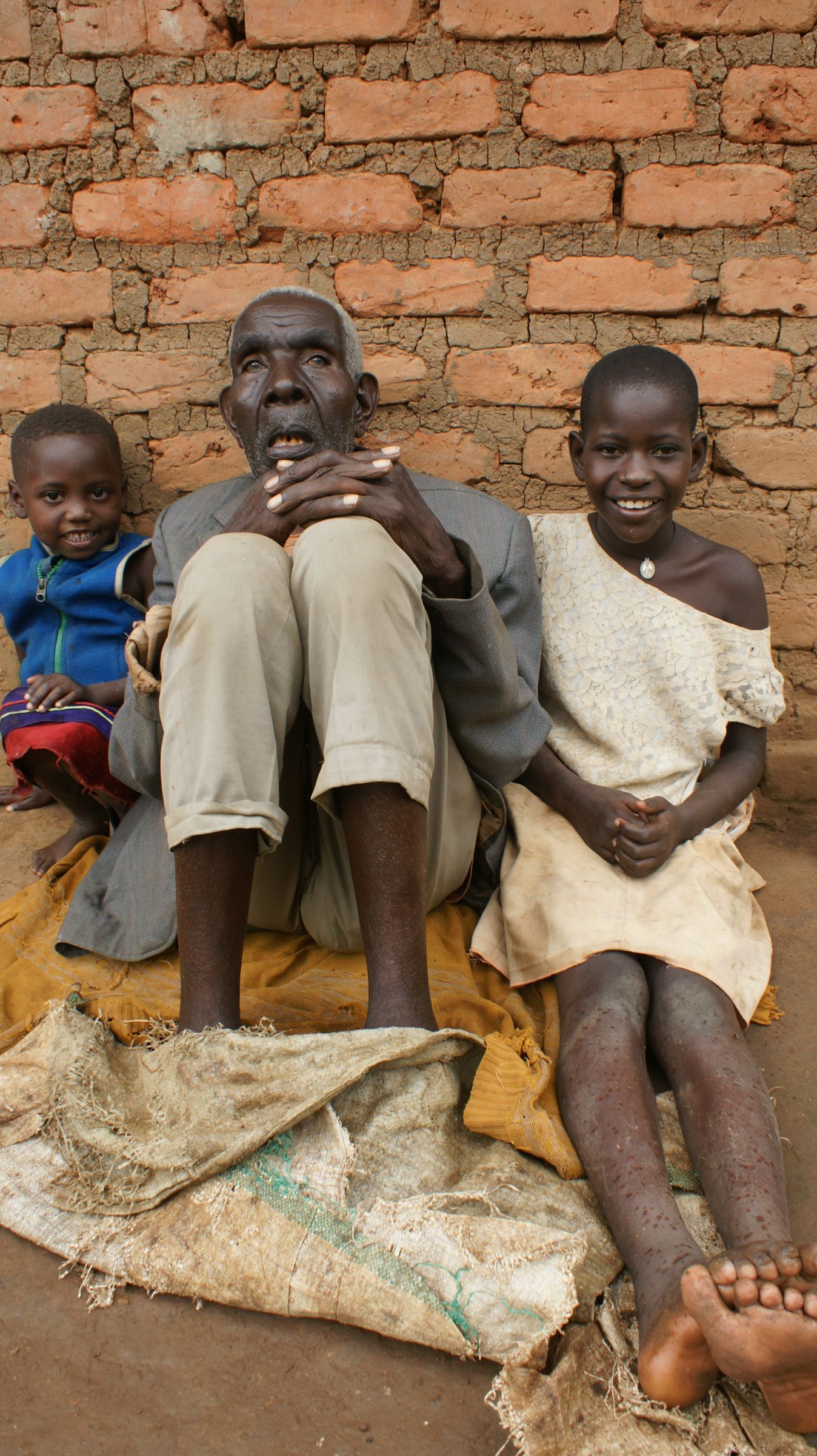 An elderly person and two children sit on a sack cloth in front of a brick wall. All are smiling.