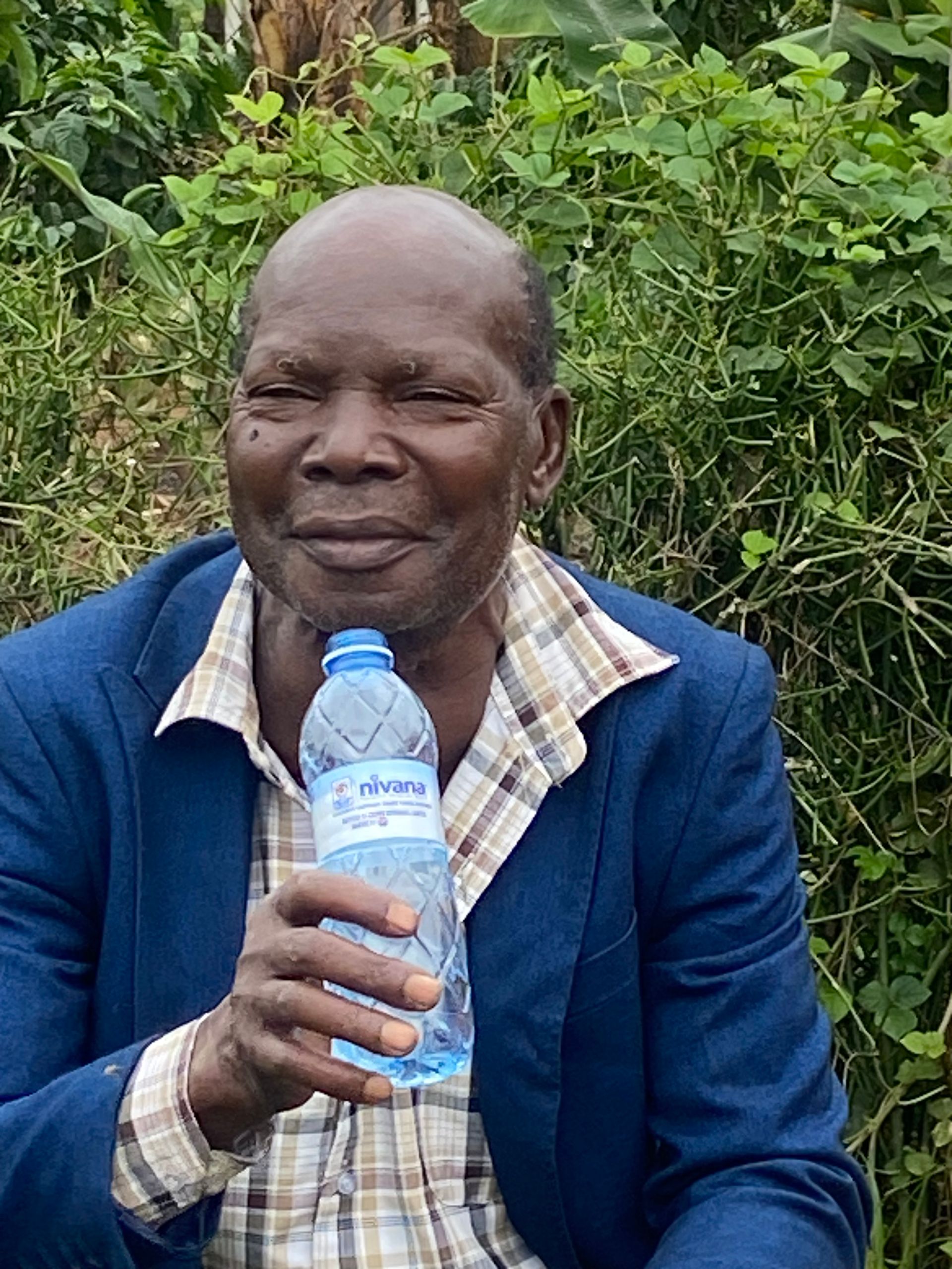 Older person holding a water bottle, wearing a blue blazer and plaid shirt, outdoors.