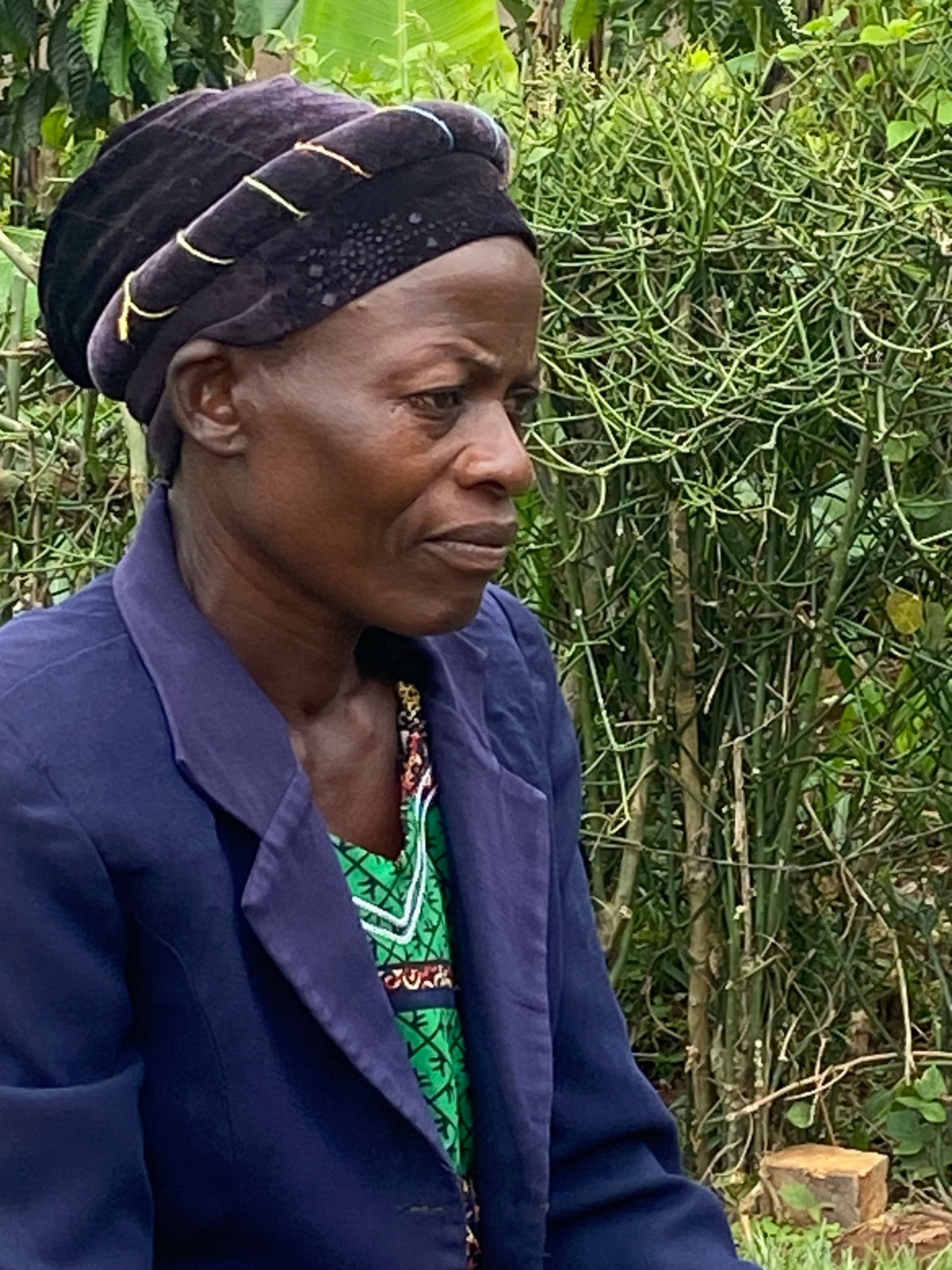 Woman wearing a black head covering and blue jacket, looking to the side, outside near greenery.