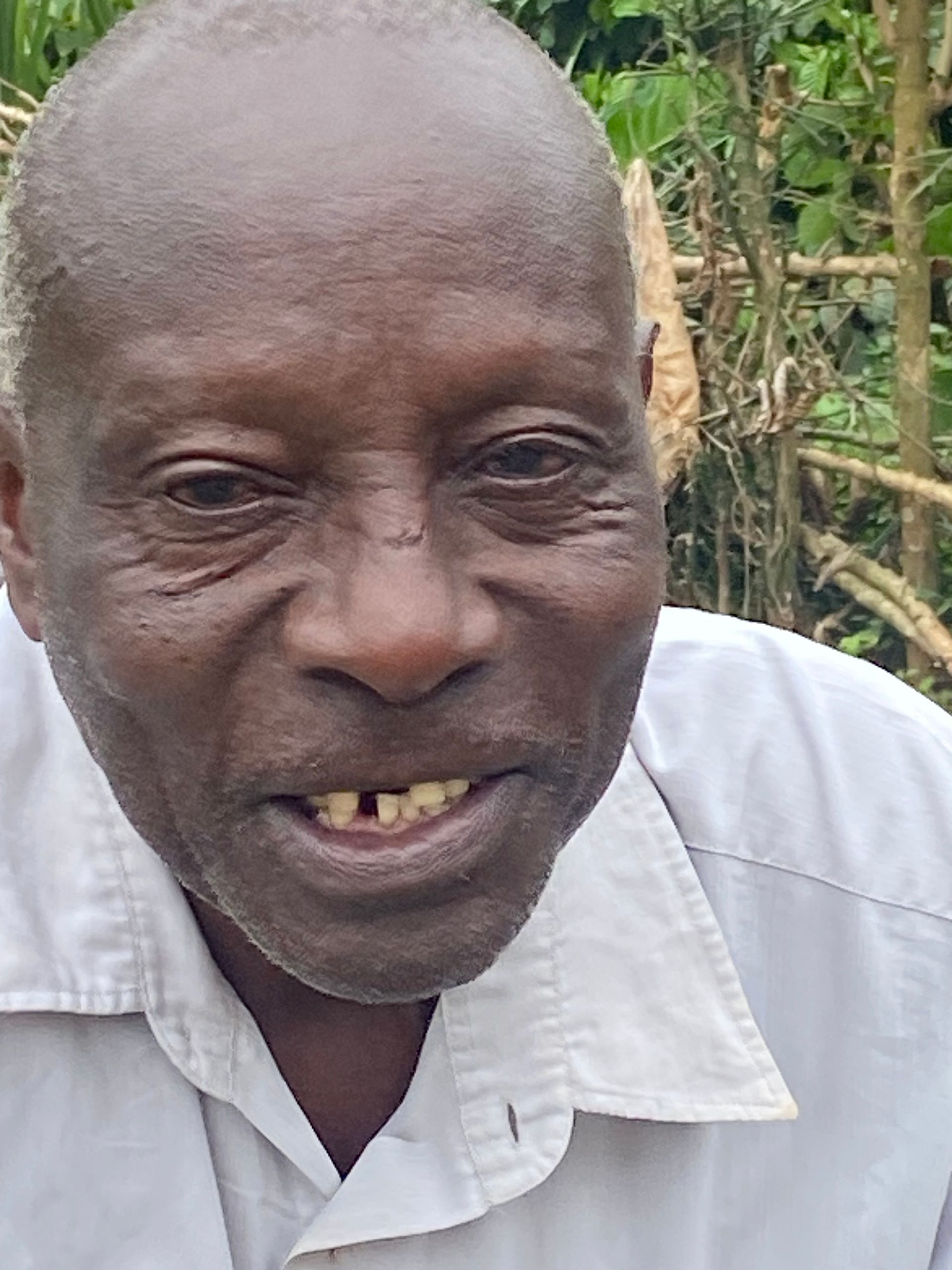 Close-up of a smiling person with dark skin, wearing a white collared shirt, outdoors.