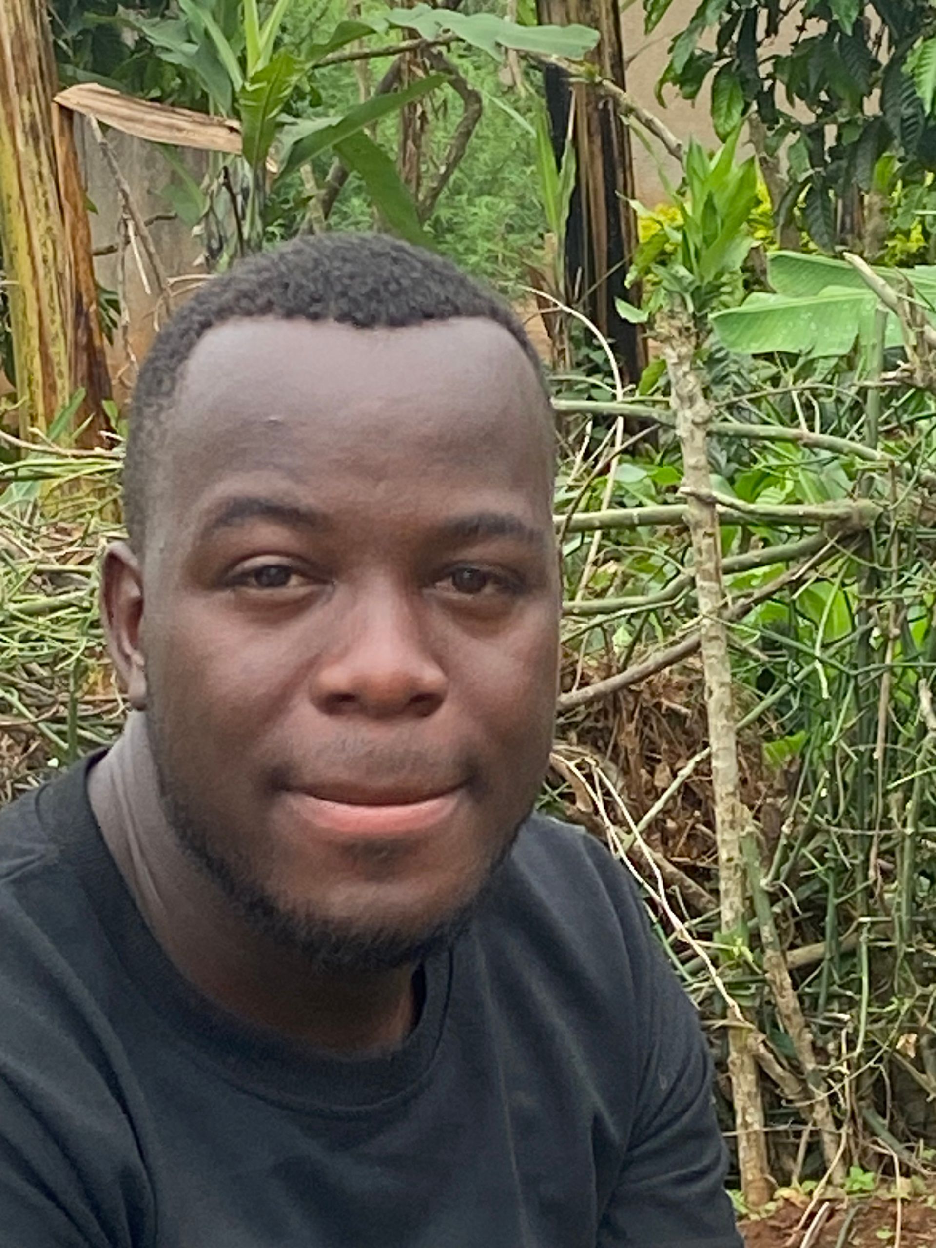 Man in black shirt smiles outdoors, foliage in background.