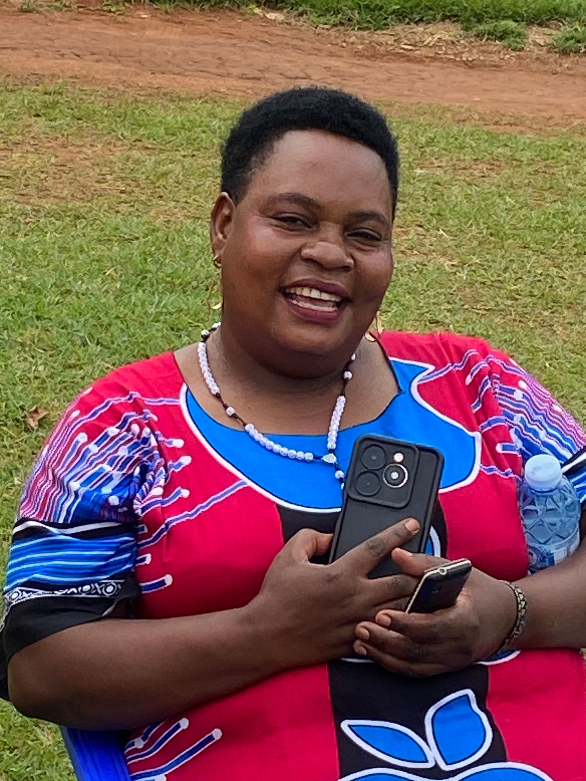 Woman in colorful dress smiling, holding phone. Sitting outside, green background.