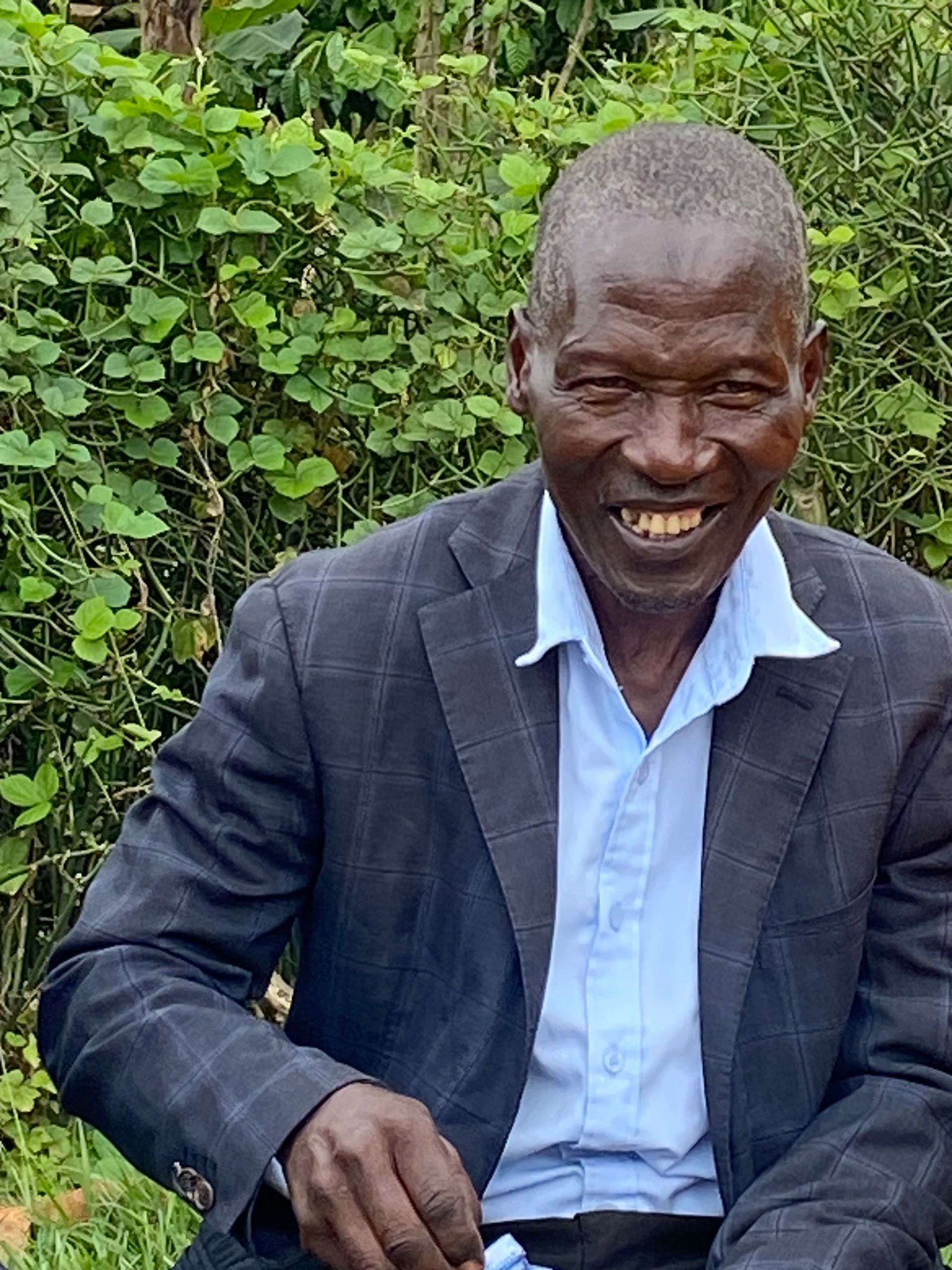 Man in dark jacket and light blue shirt smiles, seated outdoors near greenery.