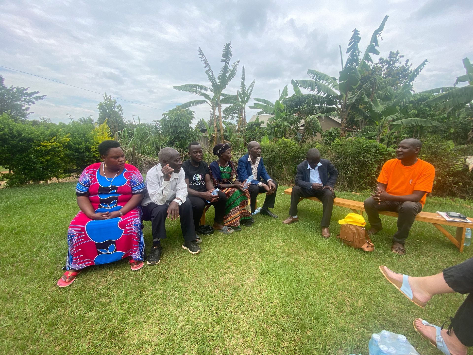 Group of people sitting on grass outdoors, possibly discussing; cloudy sky, green foliage.