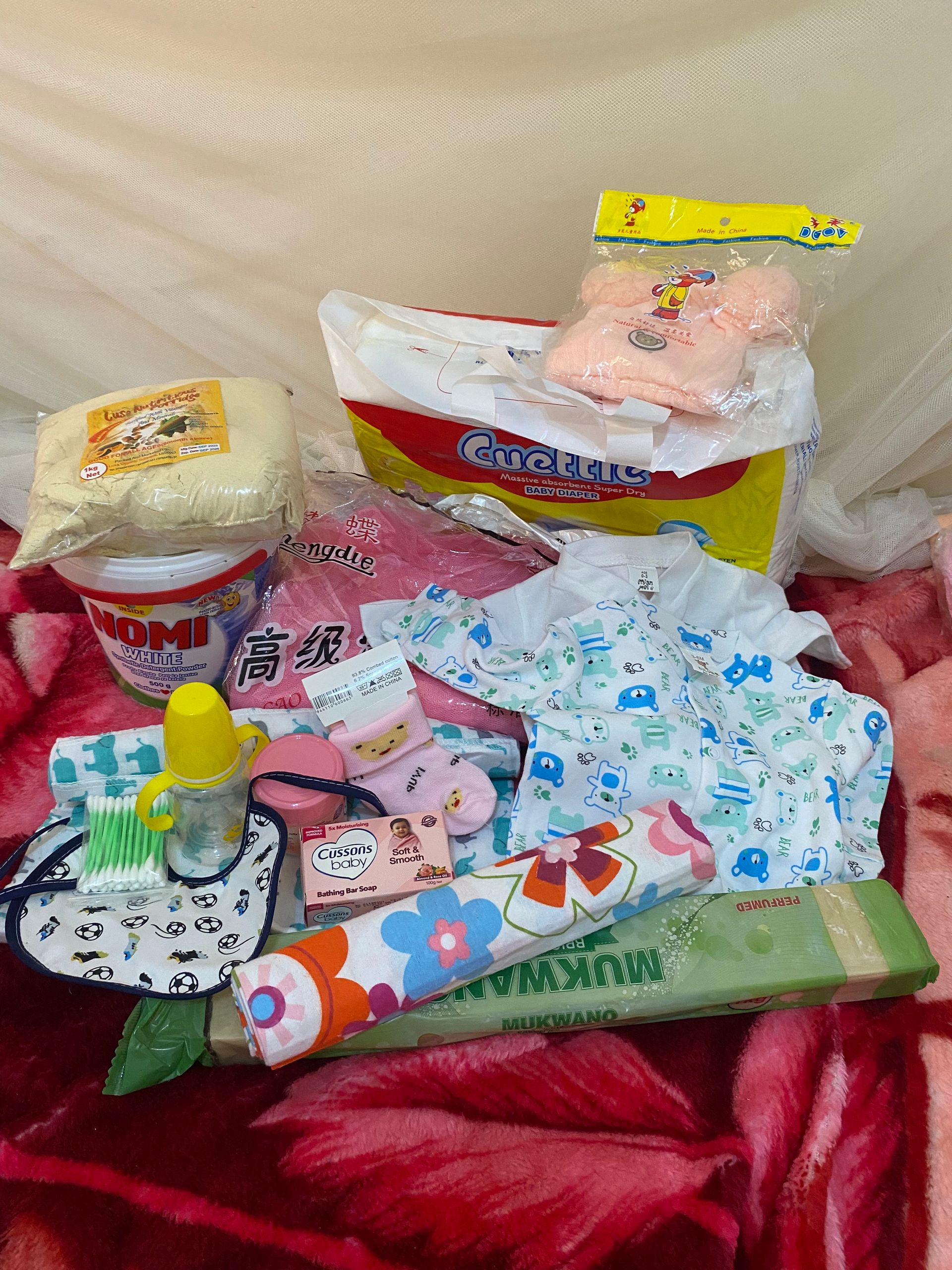 Baby items: diapers, clothes, bottle, bib, booties, and other supplies on a red blanket.