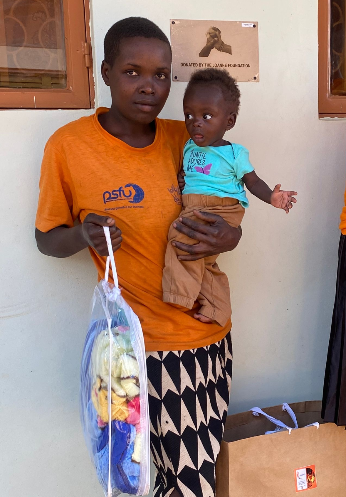 Woman in orange shirt holds a baby and a bag of items. Building background.