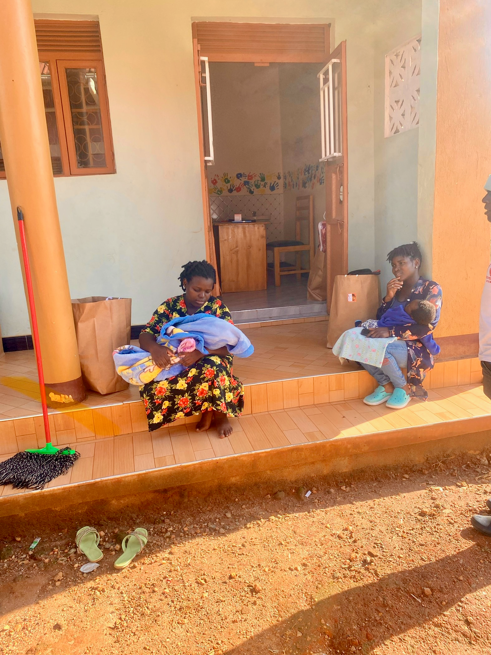 Two women outside a building: one holding a baby, the other seated. Brown ground and a partially open door.