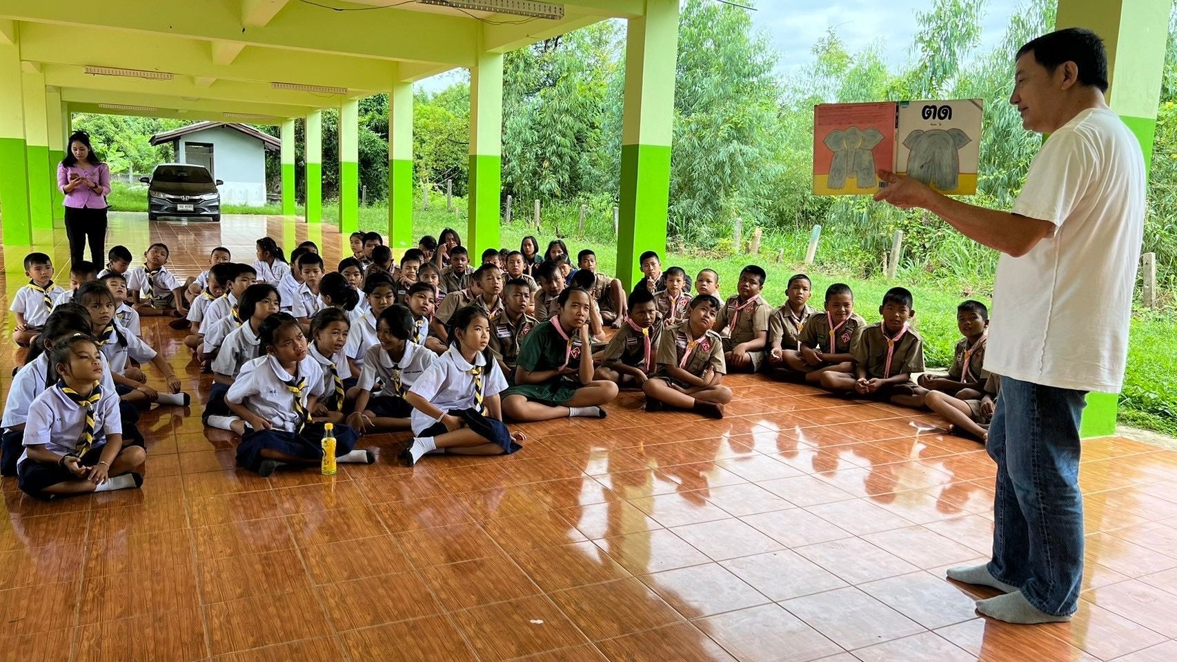 Teacher reading to students seated on floor in an outdoor school setting. Bright colors, learning activity.
