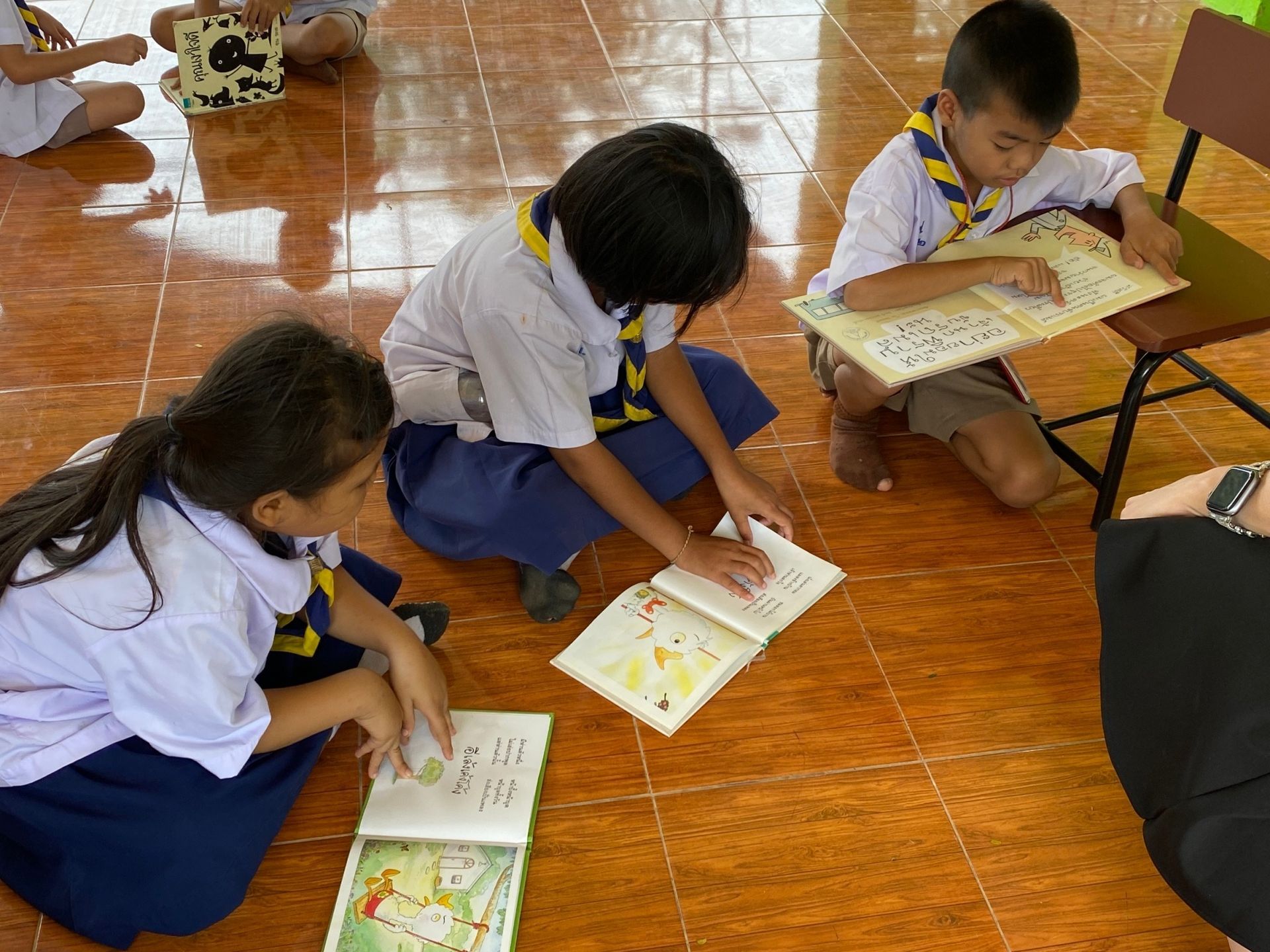 Three children in uniform reading books on the floor. One boy reads Braille.
