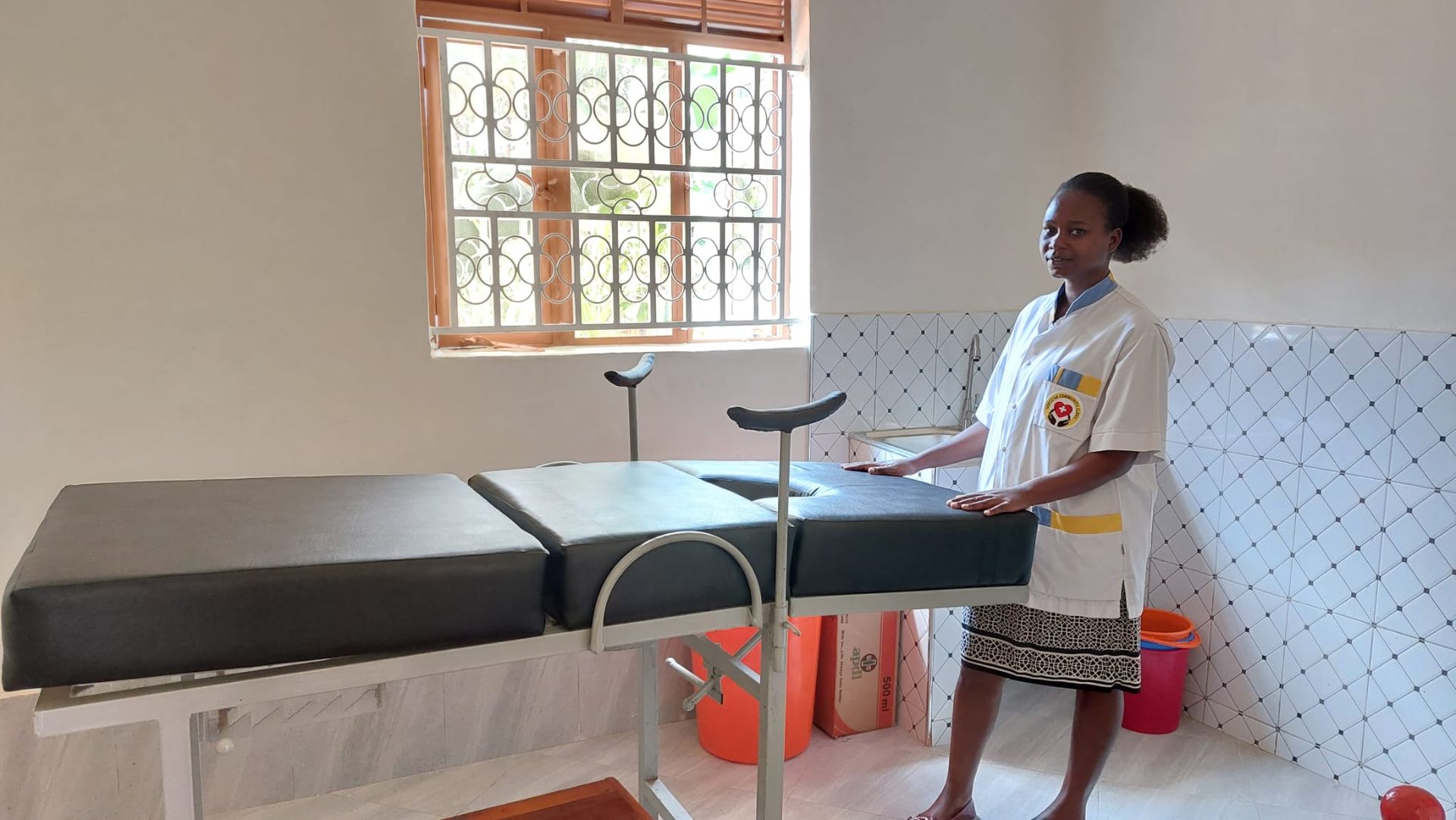 Woman in medical scrubs stands near a medical examination table in a clinic room.