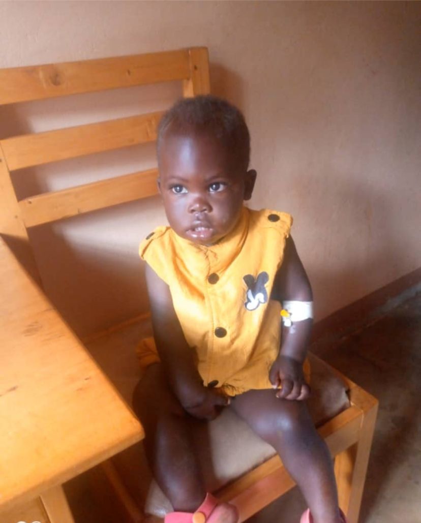 Child in yellow shirt sits on a wooden chair, looking forward with a serious expression.