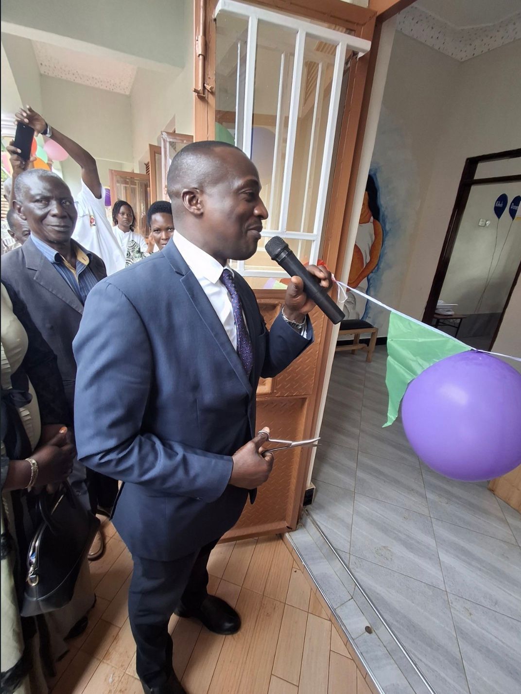 Man in suit speaking into a microphone near a doorway, a purple balloon visible.