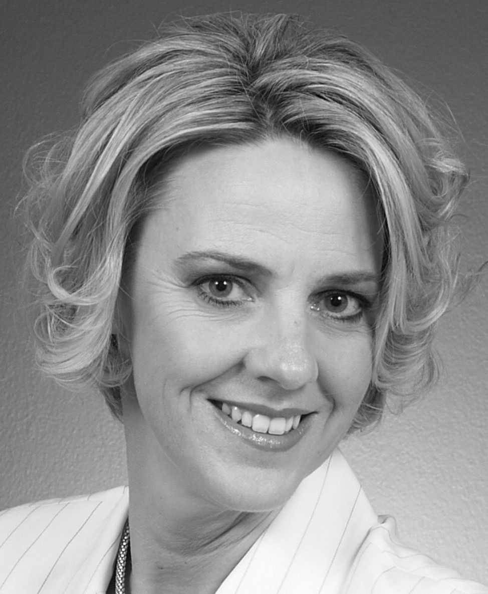 Woman with short blonde hair smiles, wearing a light-colored blazer.