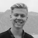 Young man with blonde hair smiles broadly, background shows a mountain and water.
