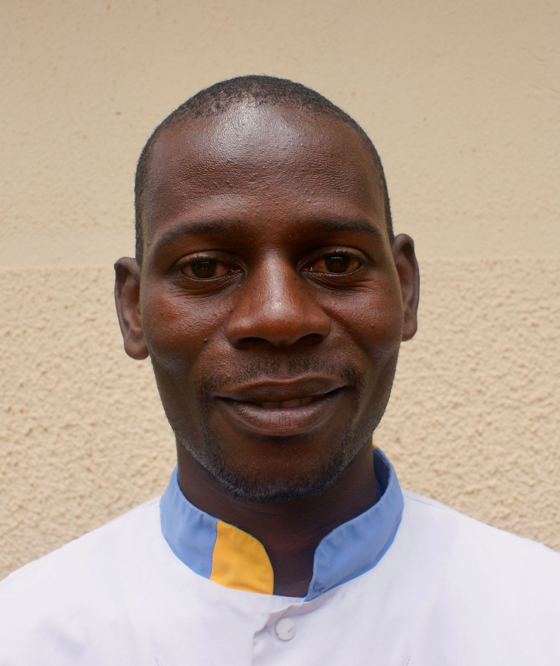 Man with short hair wearing a white uniform with blue and yellow collar, smiling.