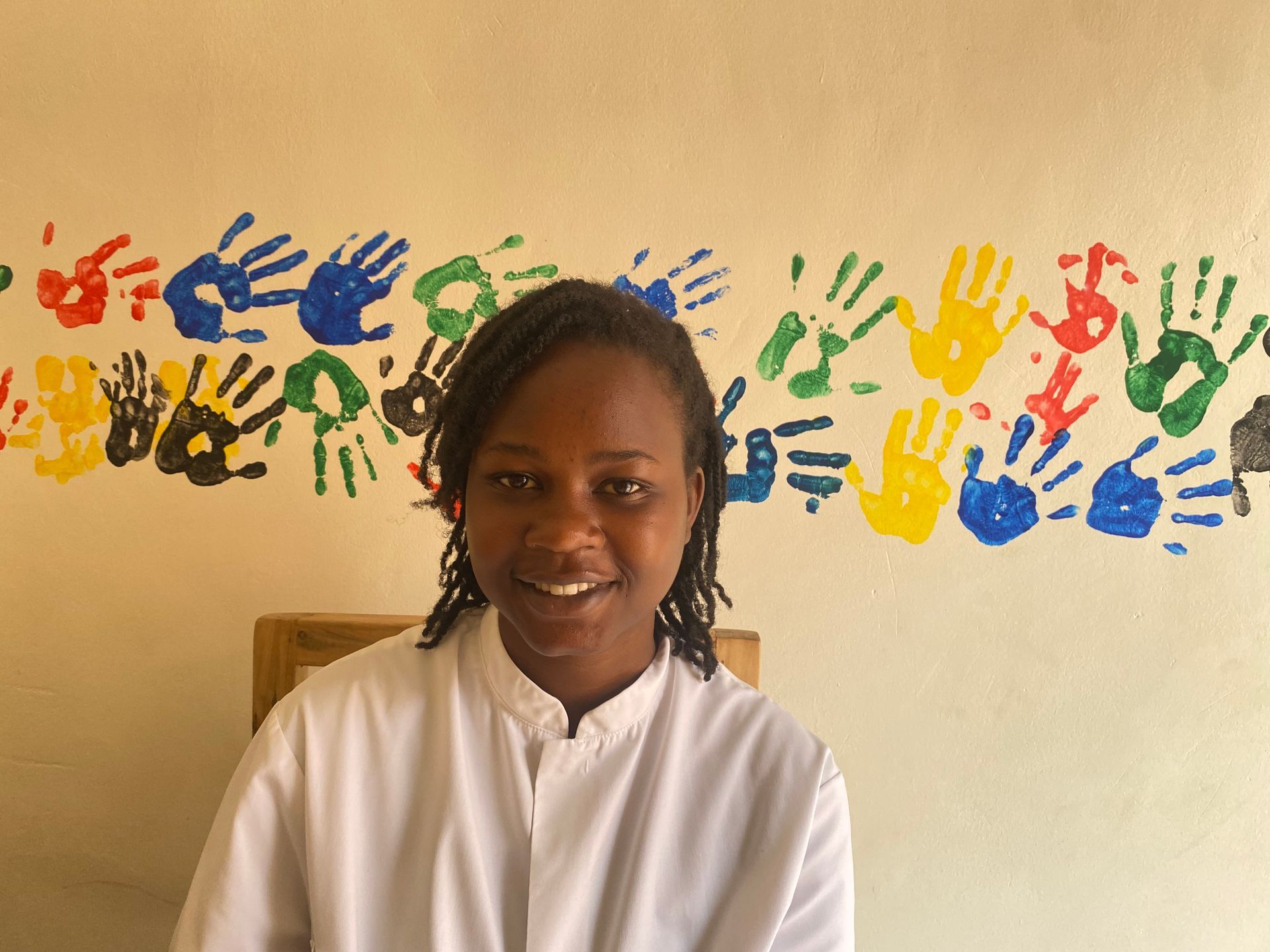 Woman in white shirt smiles, sitting before a wall decorated with colorful handprints.