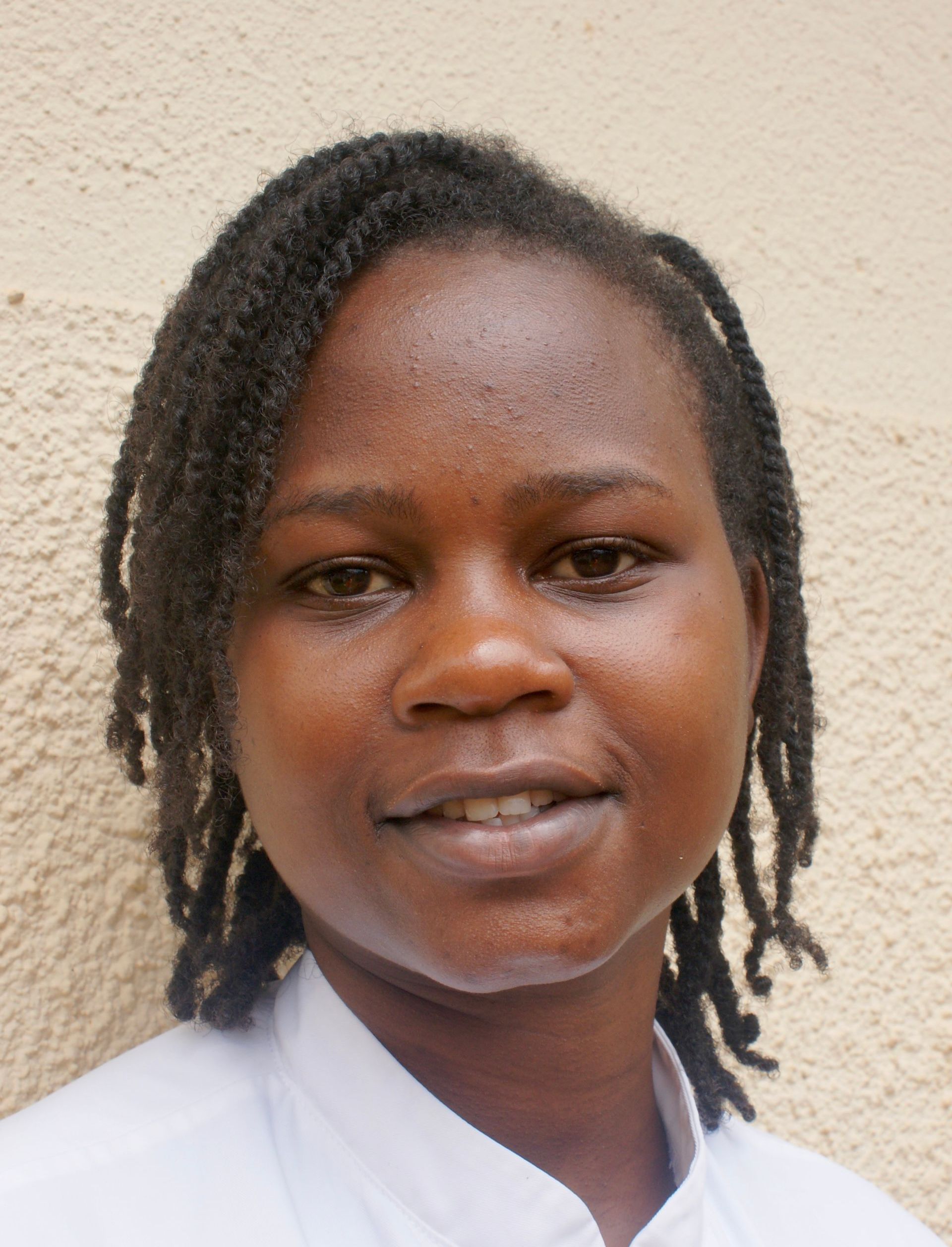 Woman with dark hair looking at the camera, wearing a white top, against a beige wall.