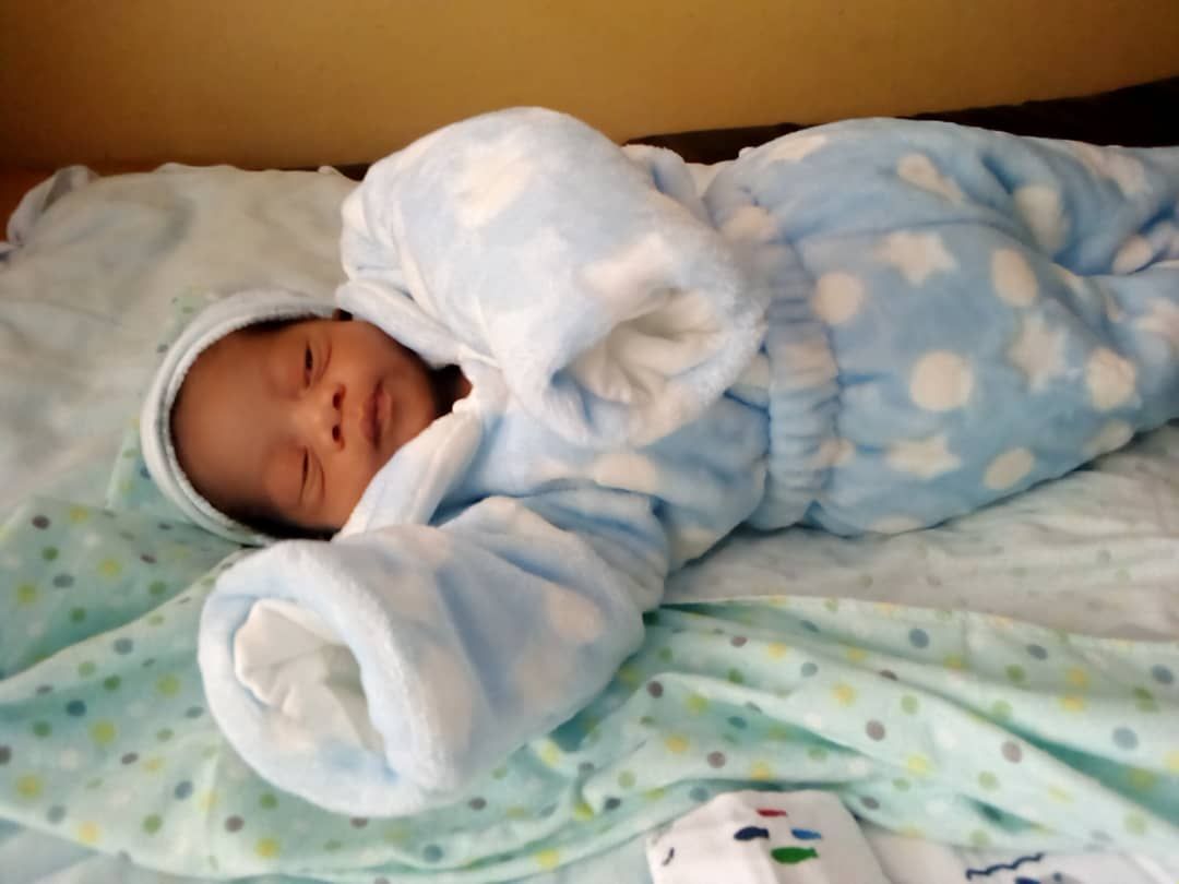 Newborn wrapped in a blue onesie with white stars, sleeping on a patterned blanket.
