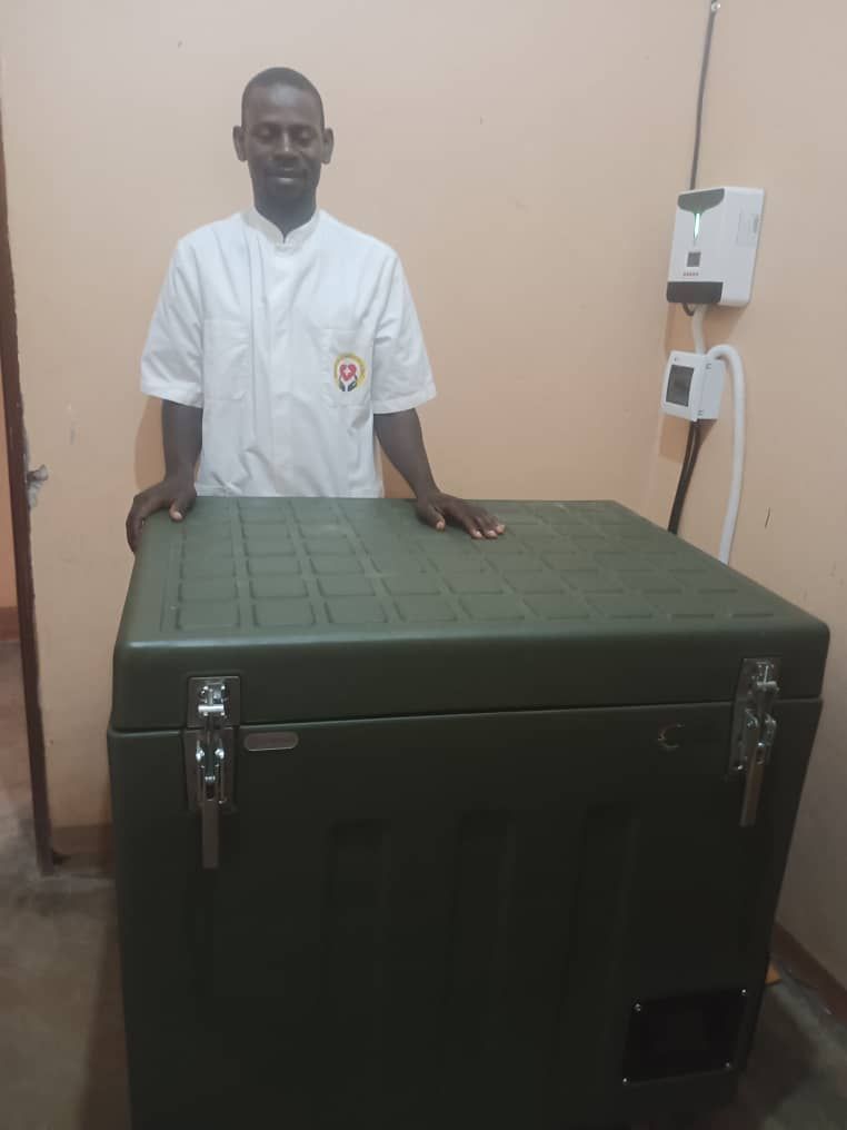Man in white coat stands next to a large green cooler in a room. Electrical device on the wall.