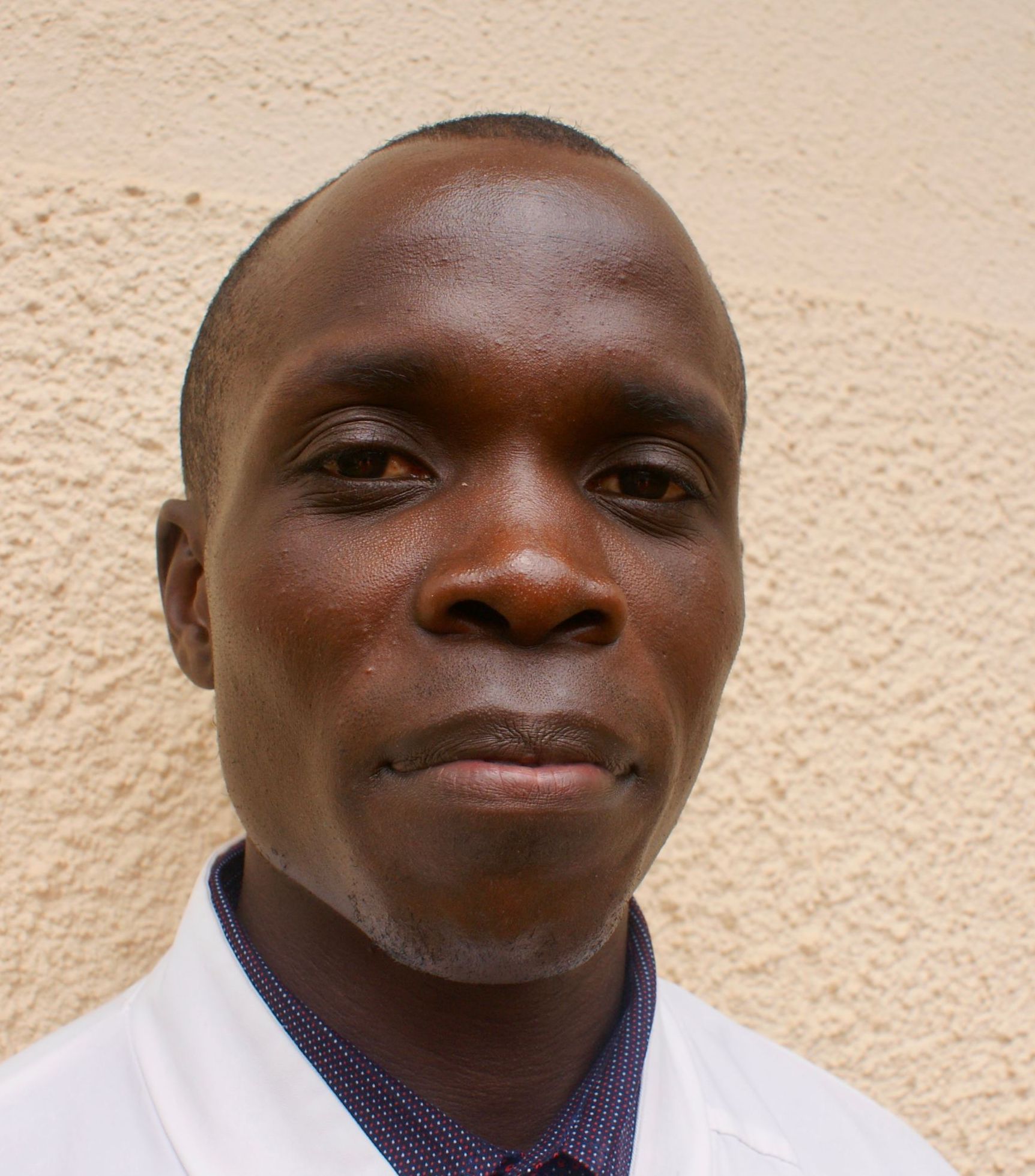 Man with short hair and dark skin wearing a white coat, looking at the camera.