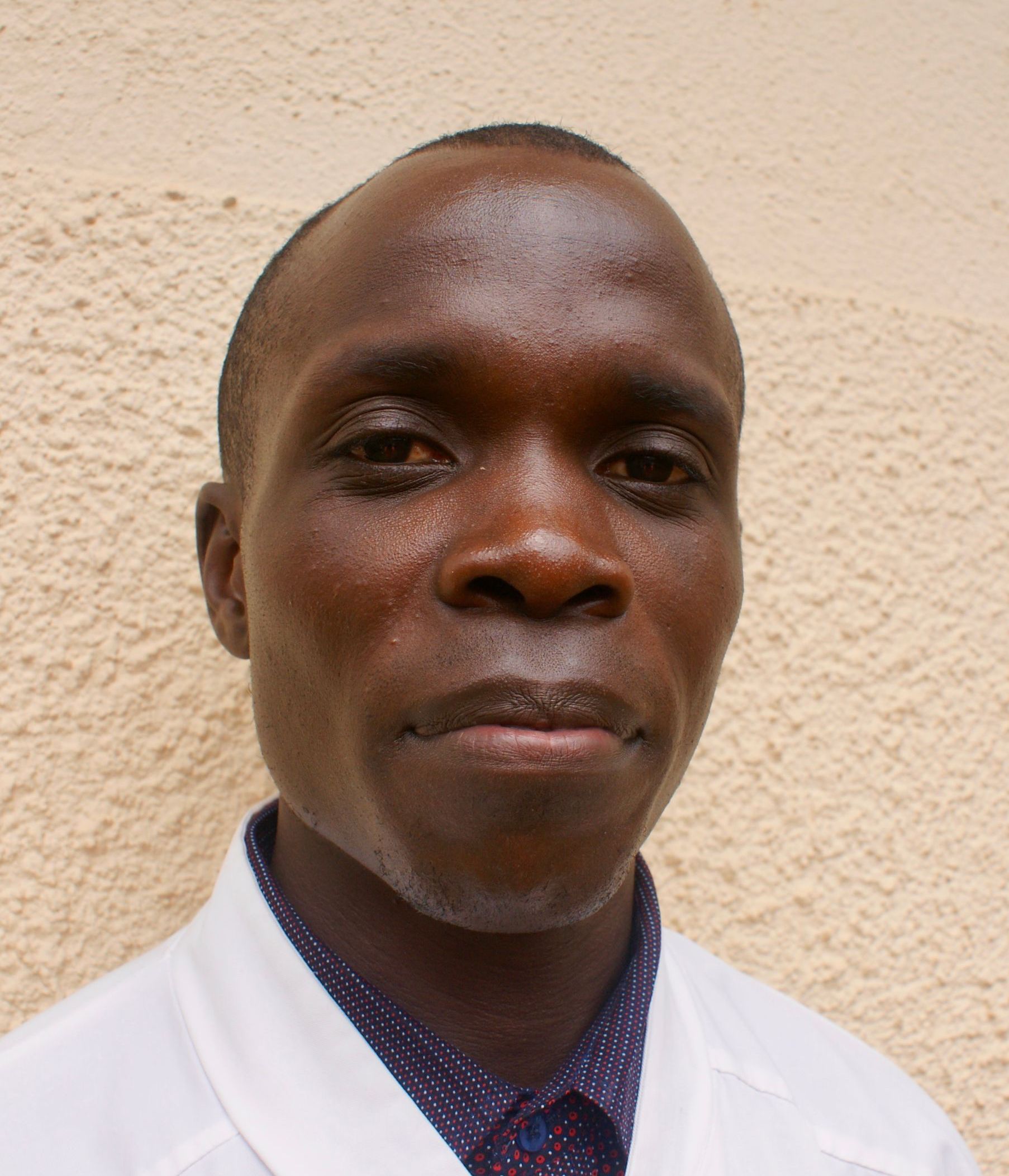 Man in white coat against a beige wall, looking directly at the camera.