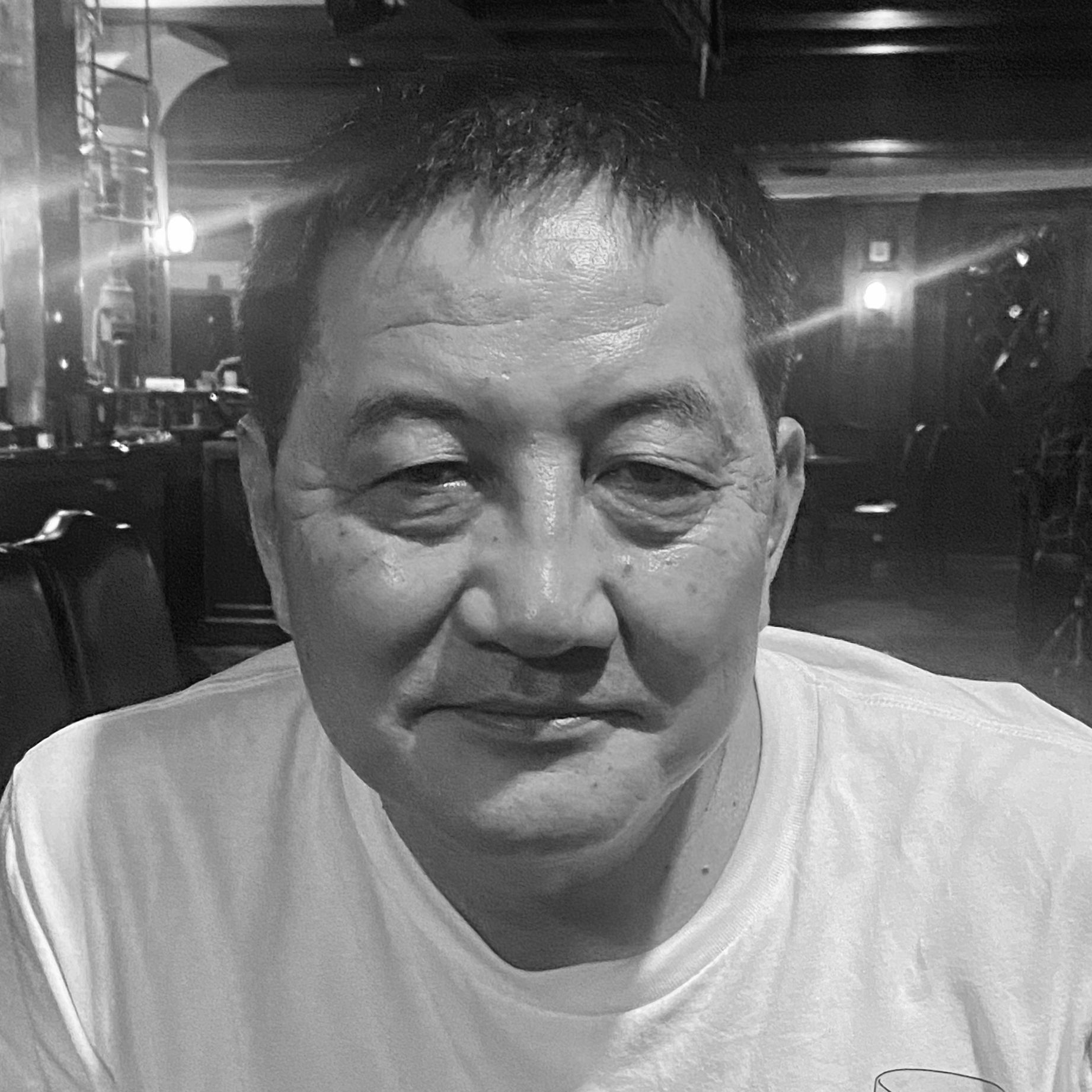 Man in a white shirt looking towards the camera, indoor, dimly lit setting.