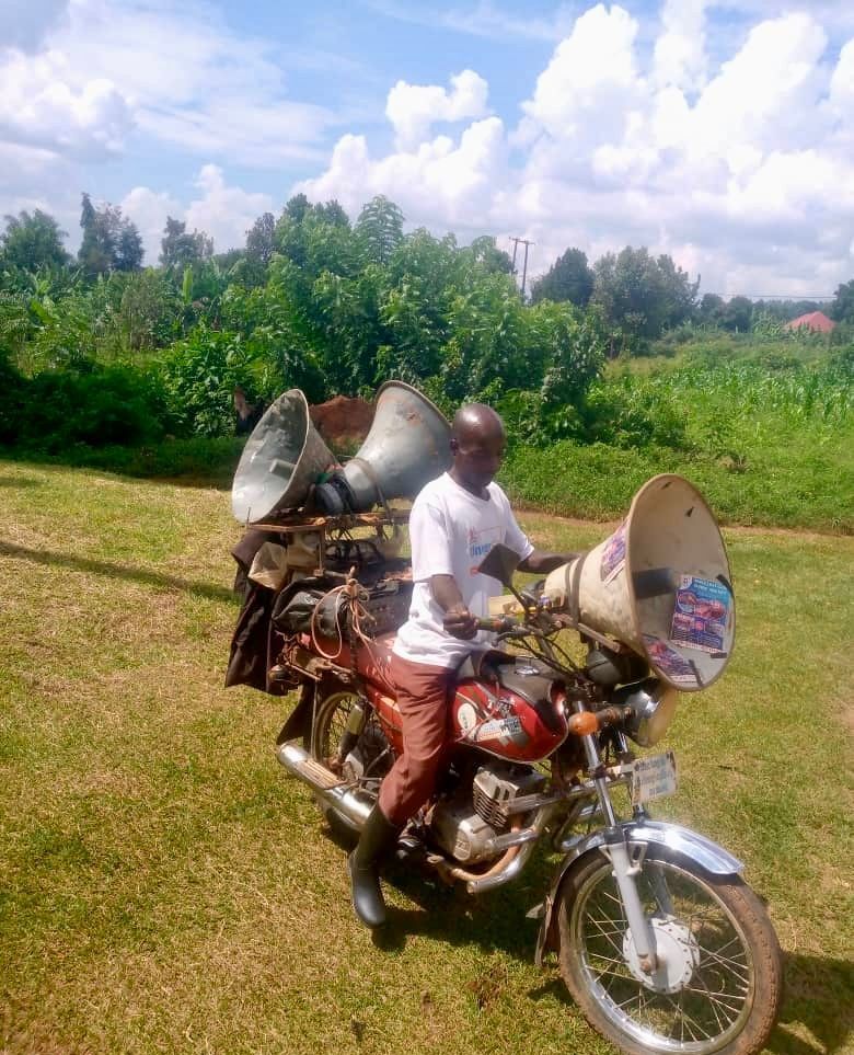 Man on motorcycle loaded with large speakers in a grassy field. Sky and trees in background.