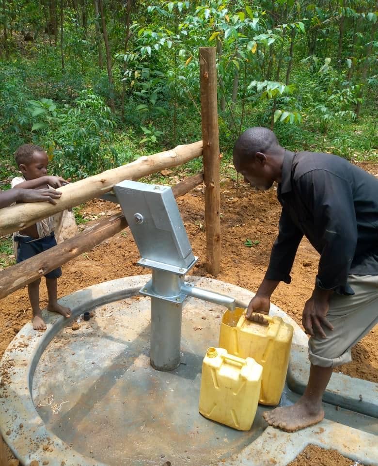 Man pumping water into yellow jugs at a well, with a child nearby, in a forested area.