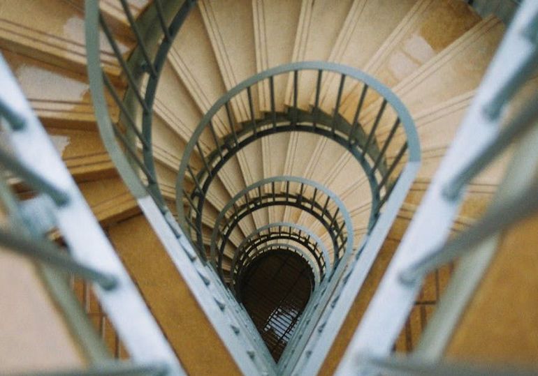 Spiral staircase with light-colored steps descending into darkness, flanked by metal railings.