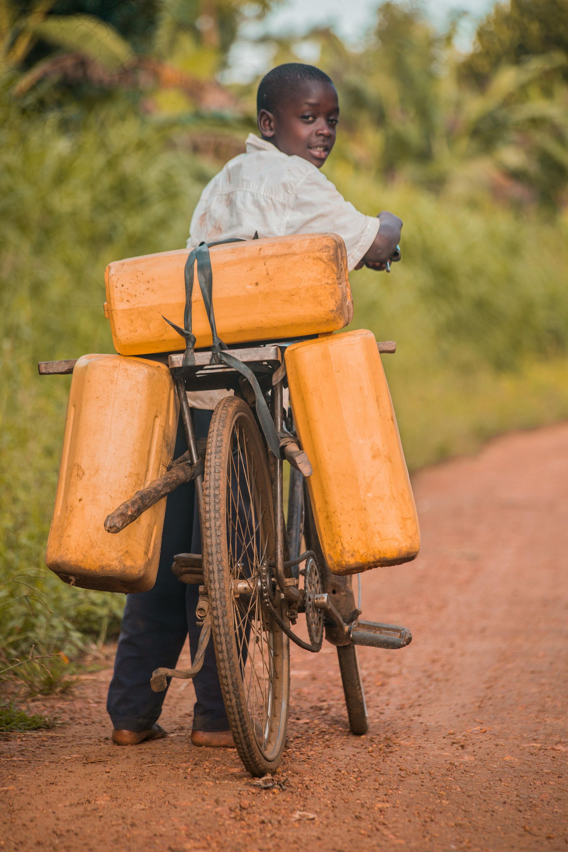 Boy on bicycle carrying water containers on dirt road.